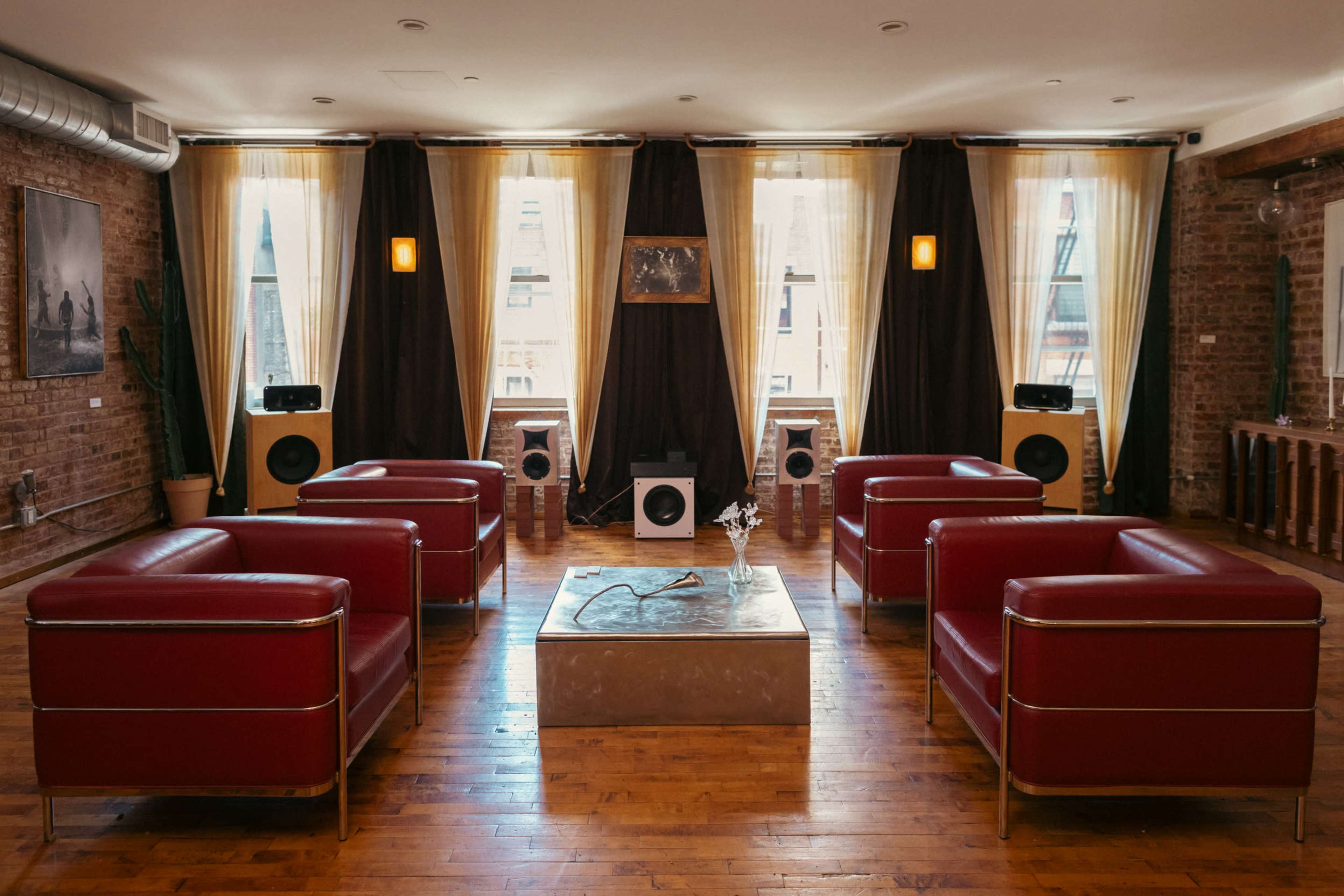 The image shows a modern lounge area with red armchairs arranged around a low, rectangular coffee table, set against a backdrop of large windows covered with flowing curtains.