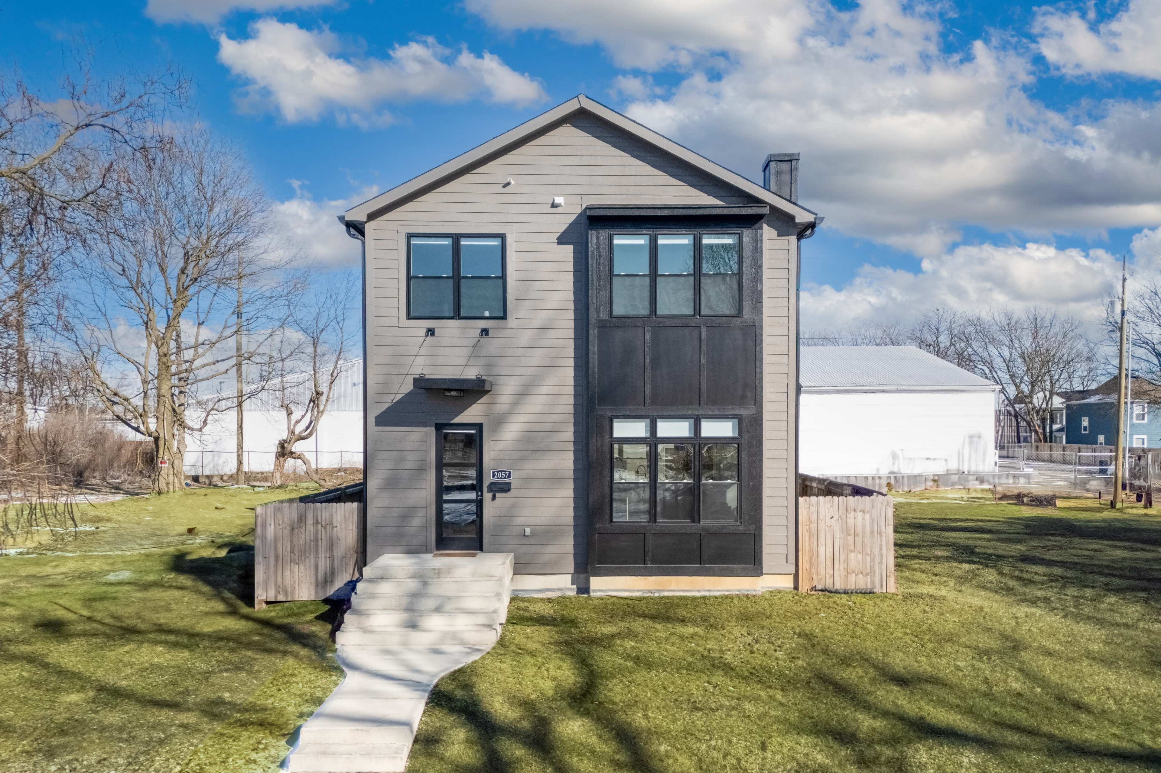 A modern two-story house with a combination of gray and black siding is set on a grassy lot, featuring a clear blue sky and scattered clouds in the background.