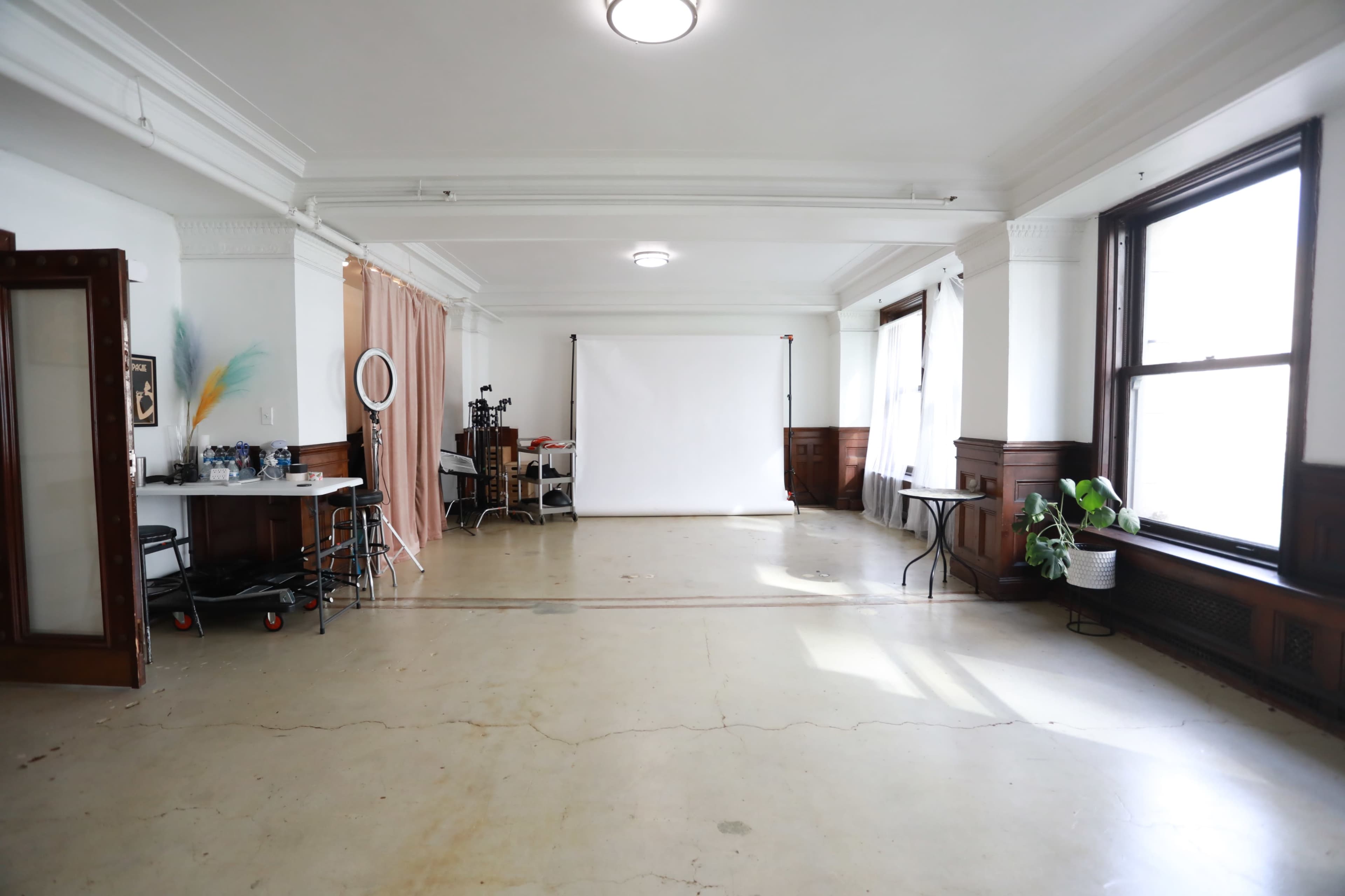 The image shows a spacious, empty studio with high ceilings, large windows, and minimalist furnishings, including a backdrop setup and some chairs.