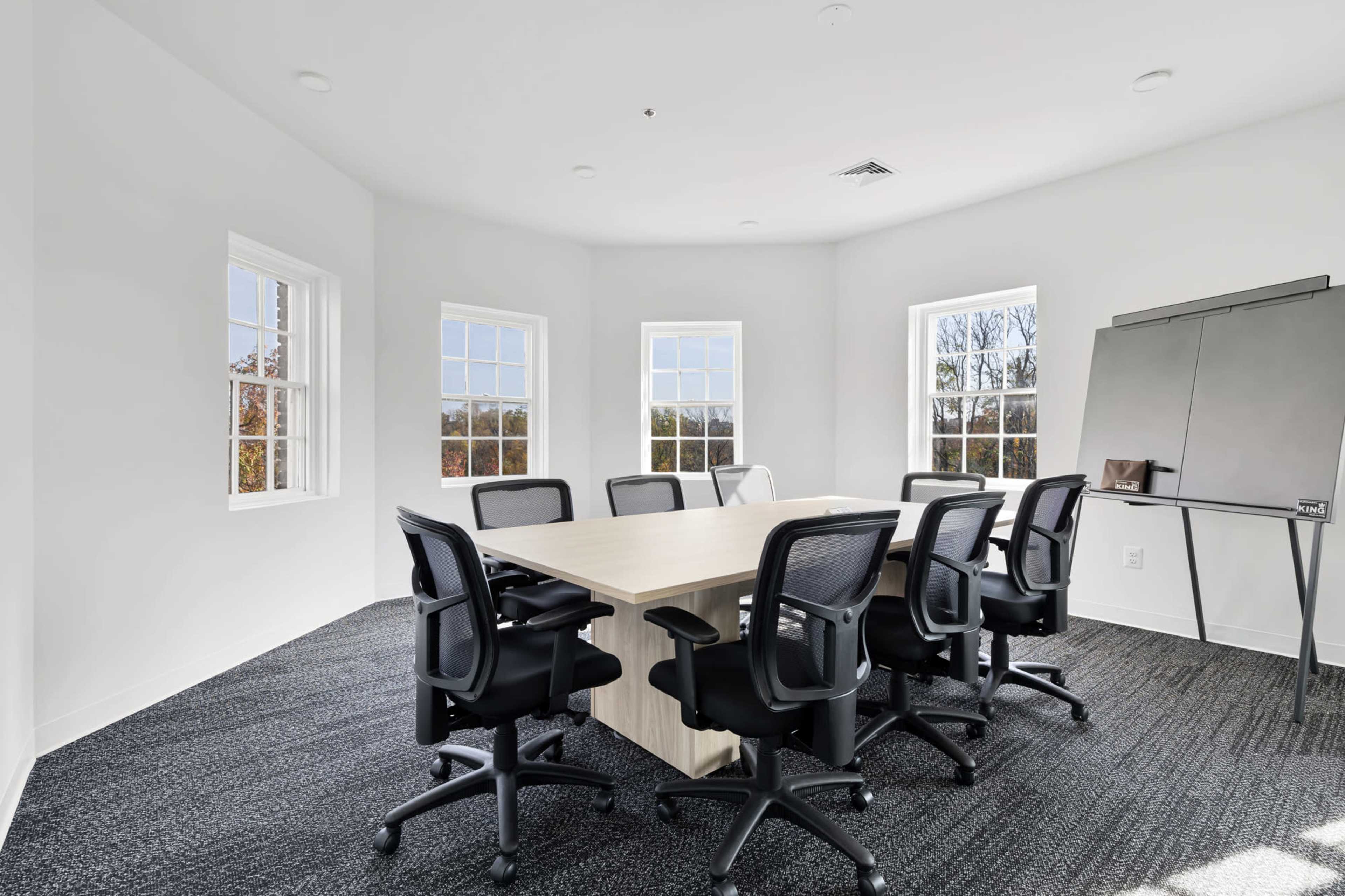A conference room features a large table surrounded by black chairs, with windows on three walls allowing natural light to enter.