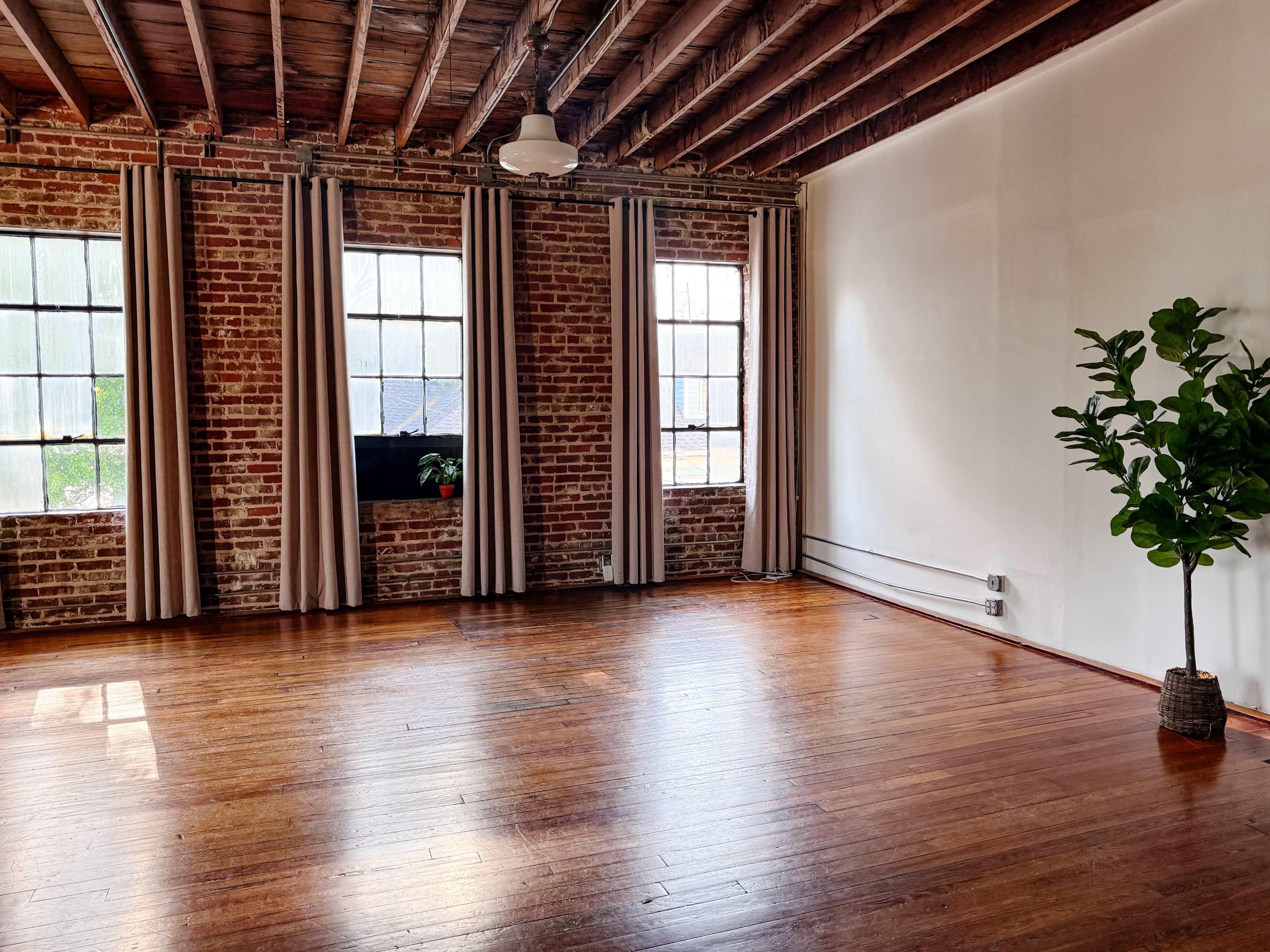 The image shows an empty room with hardwood floors, exposed brick walls, large windows with curtains, and a potted plant in the corner.