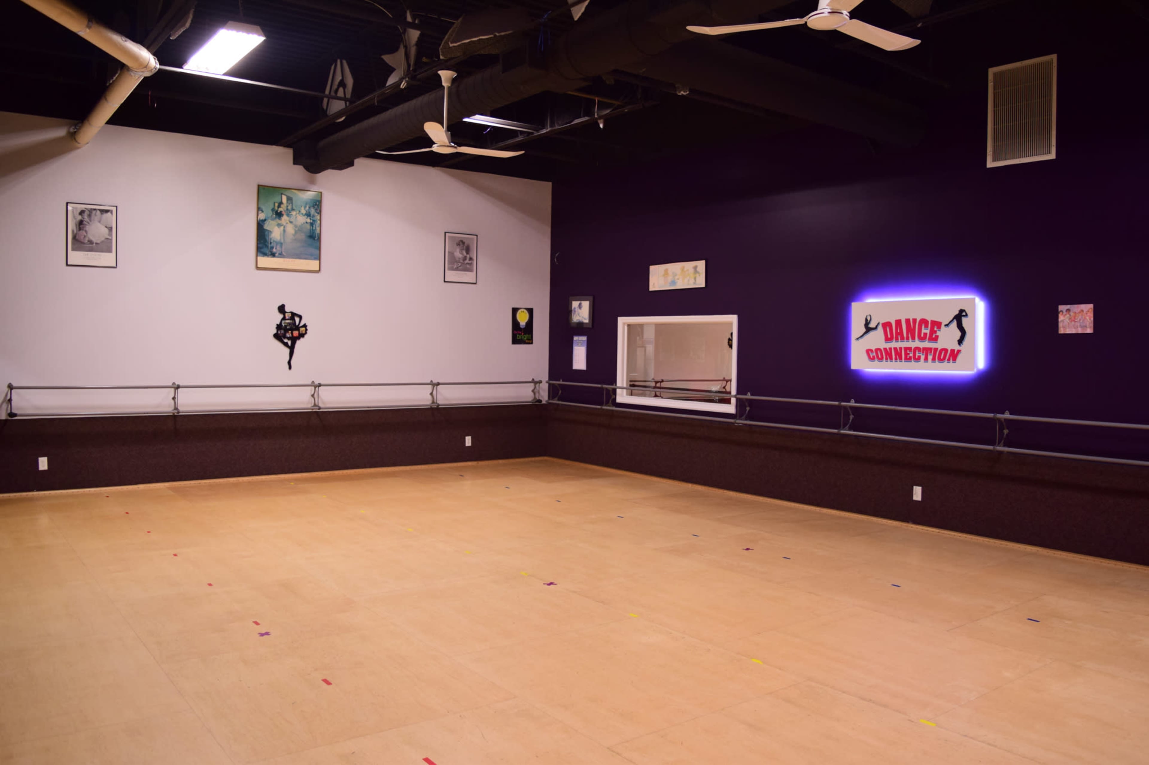 A spacious dance studio with a wooden floor, mirrored wall, and purple-painted walls, featuring the name "Dance Connection" illuminated on one side.