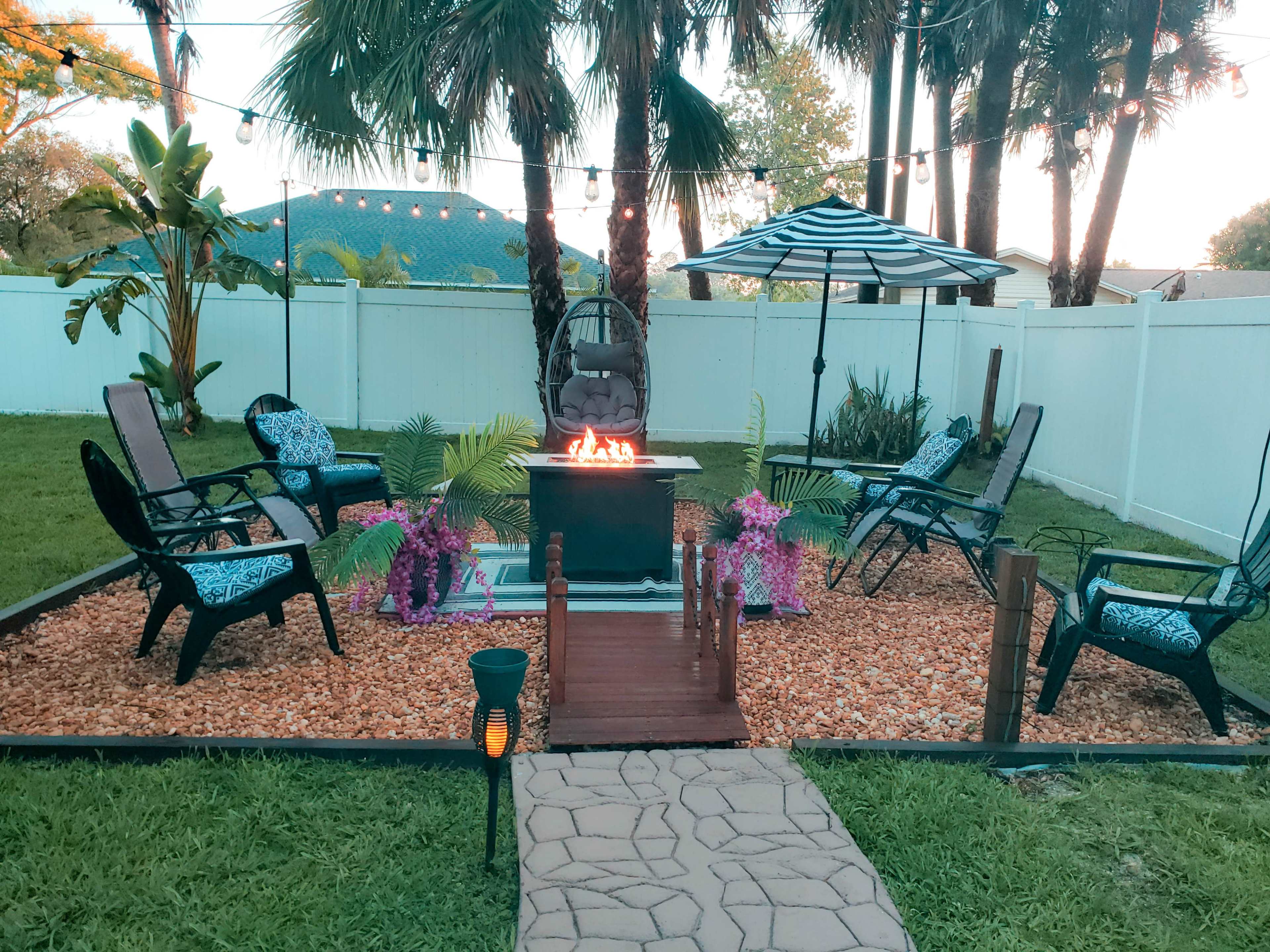 A backyard oasis features a fire pit surrounded by seating, decorative plants, and an umbrella, all set against a white fence and palm trees.