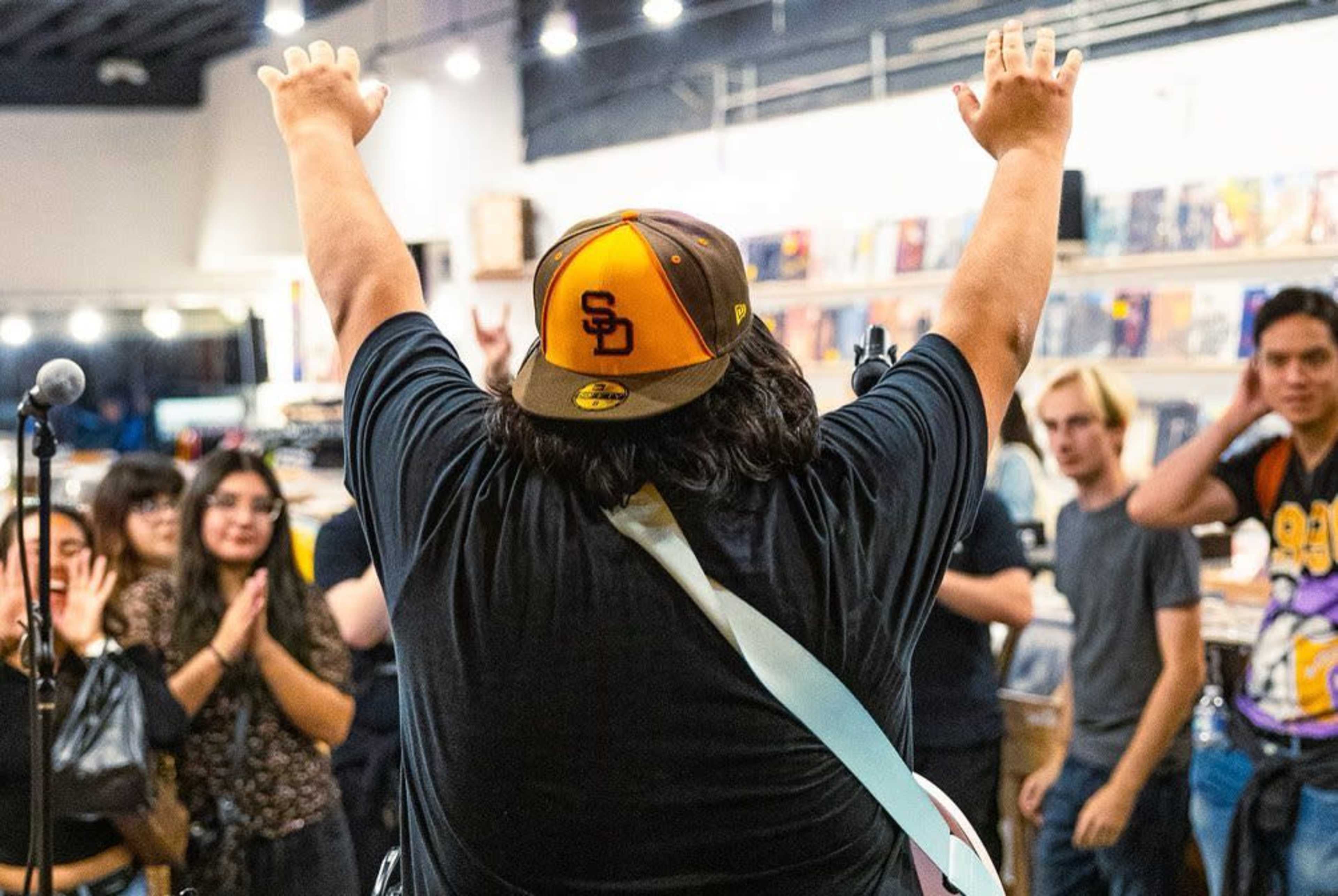 A musician stands with arms raised, performing in front of a crowd in a record store.