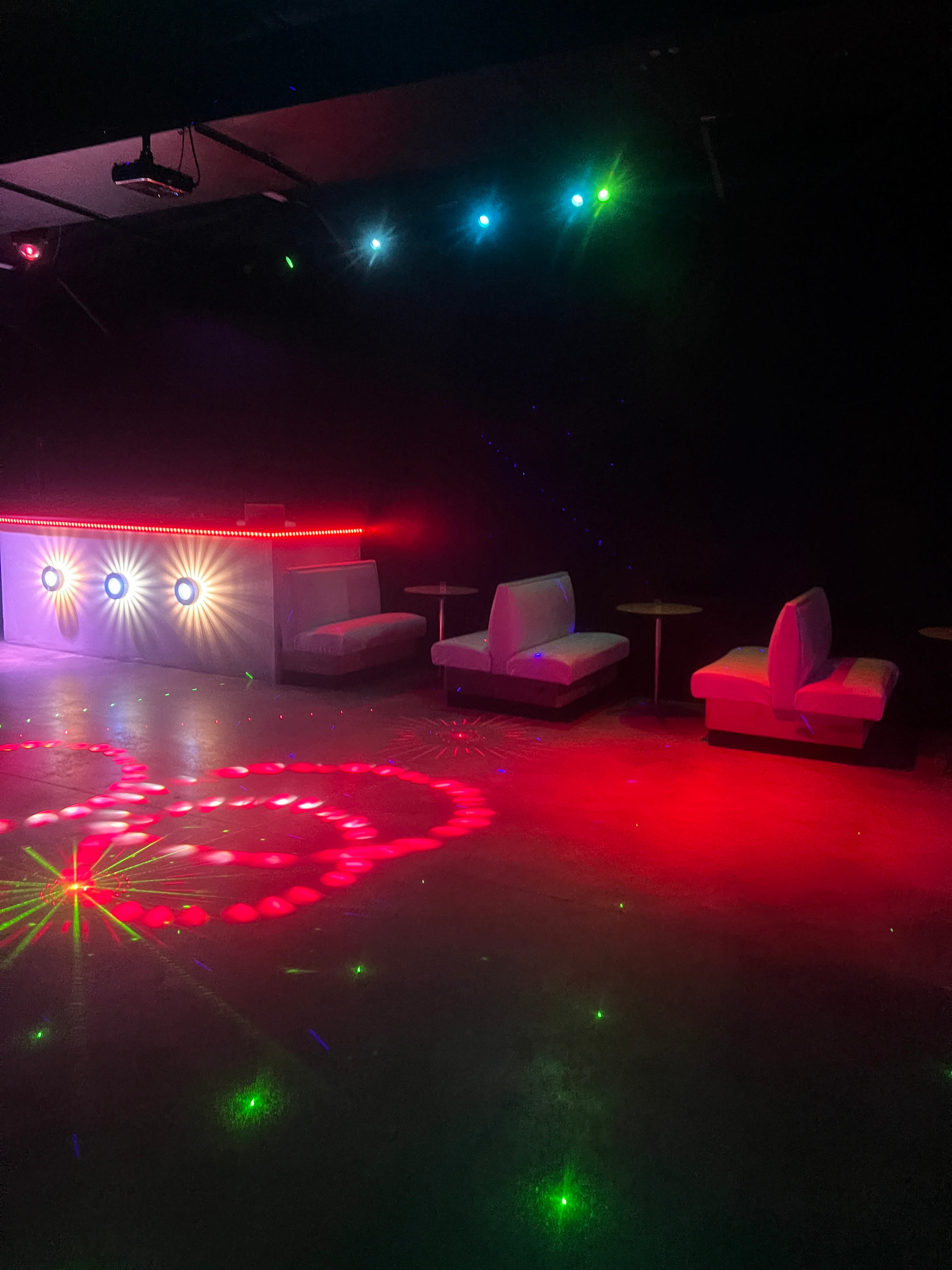 The image shows a dimly lit room with a bar featuring colored lights, white couches, and circular tables.