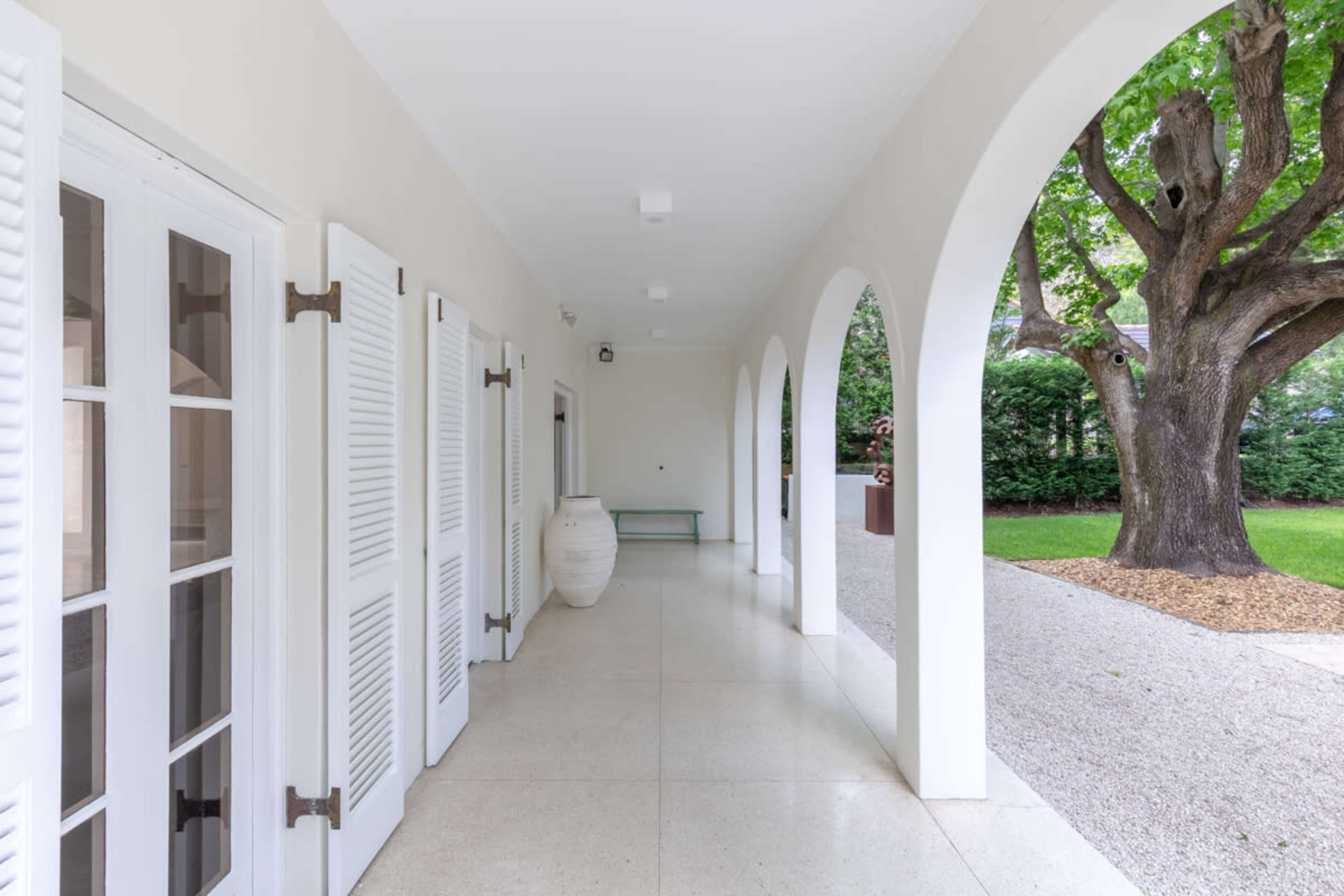 The image shows a corridor with white walls, arched openings, and a large tree visible outside.