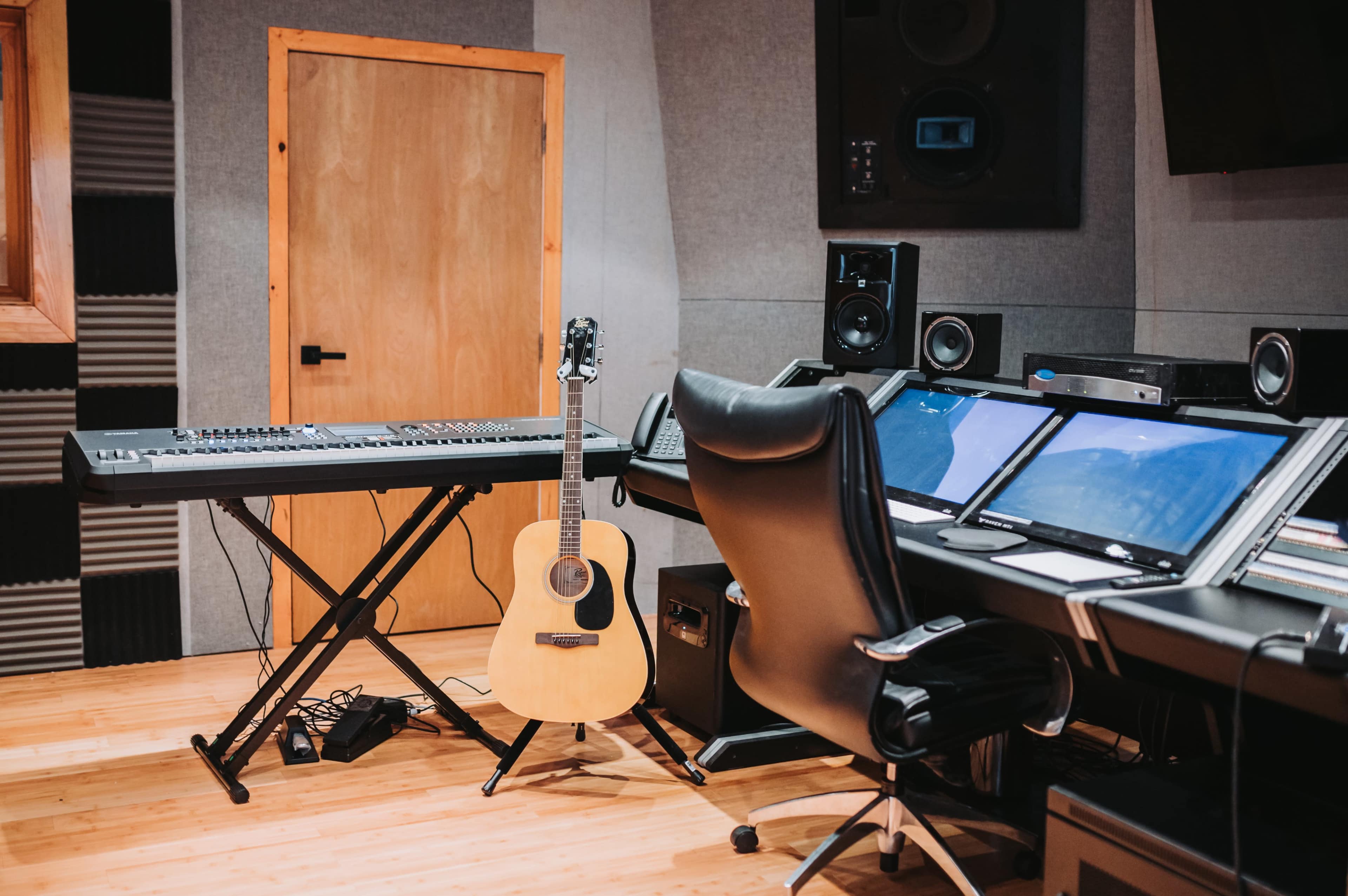 The image shows a music studio featuring a keyboard, an acoustic guitar, and a sound mixing console surrounded by acoustic panels.