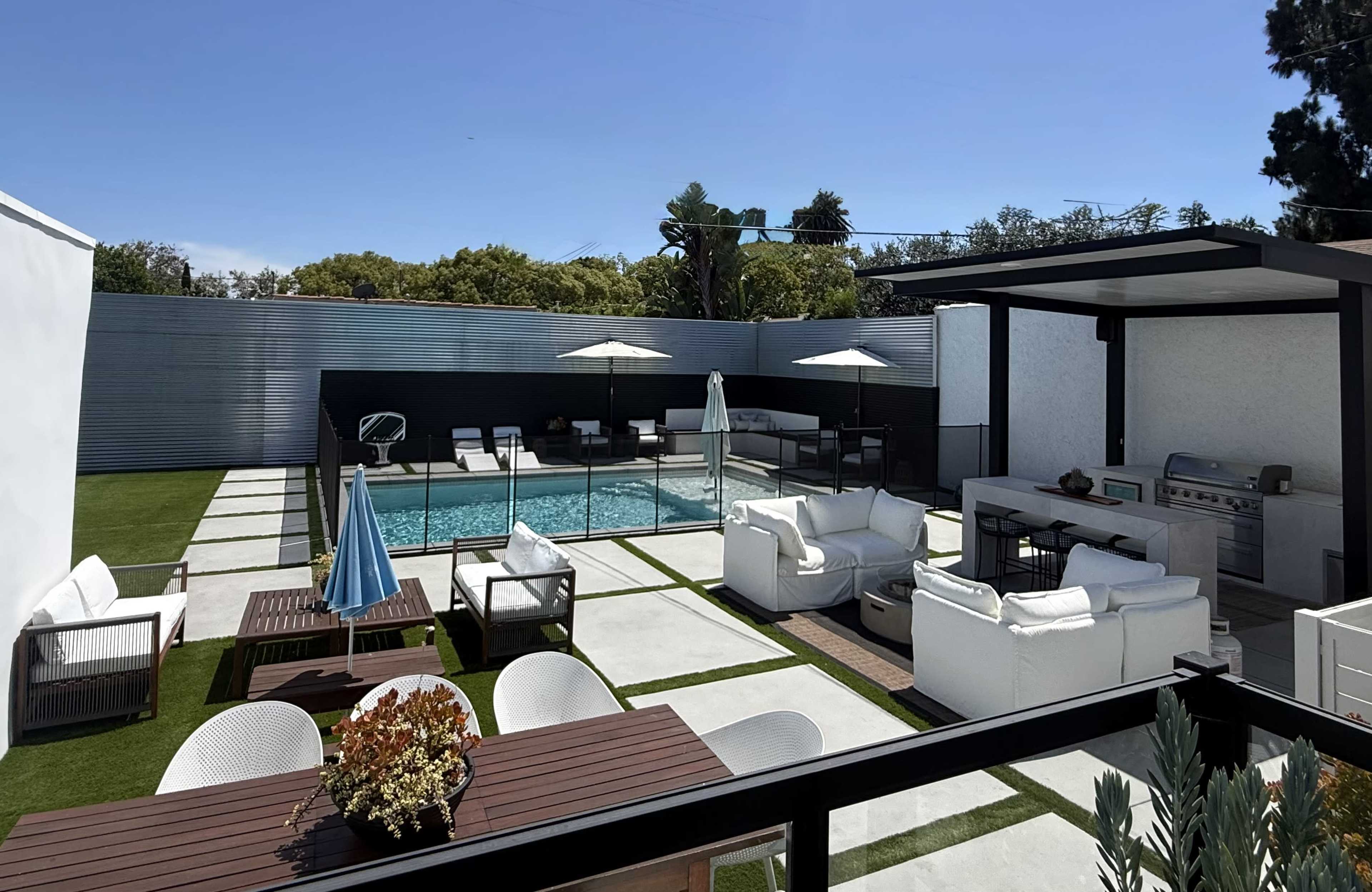 The image shows a modern outdoor patio area featuring a swimming pool surrounded by lounge chairs, a dining setup, and a charcoal grill.