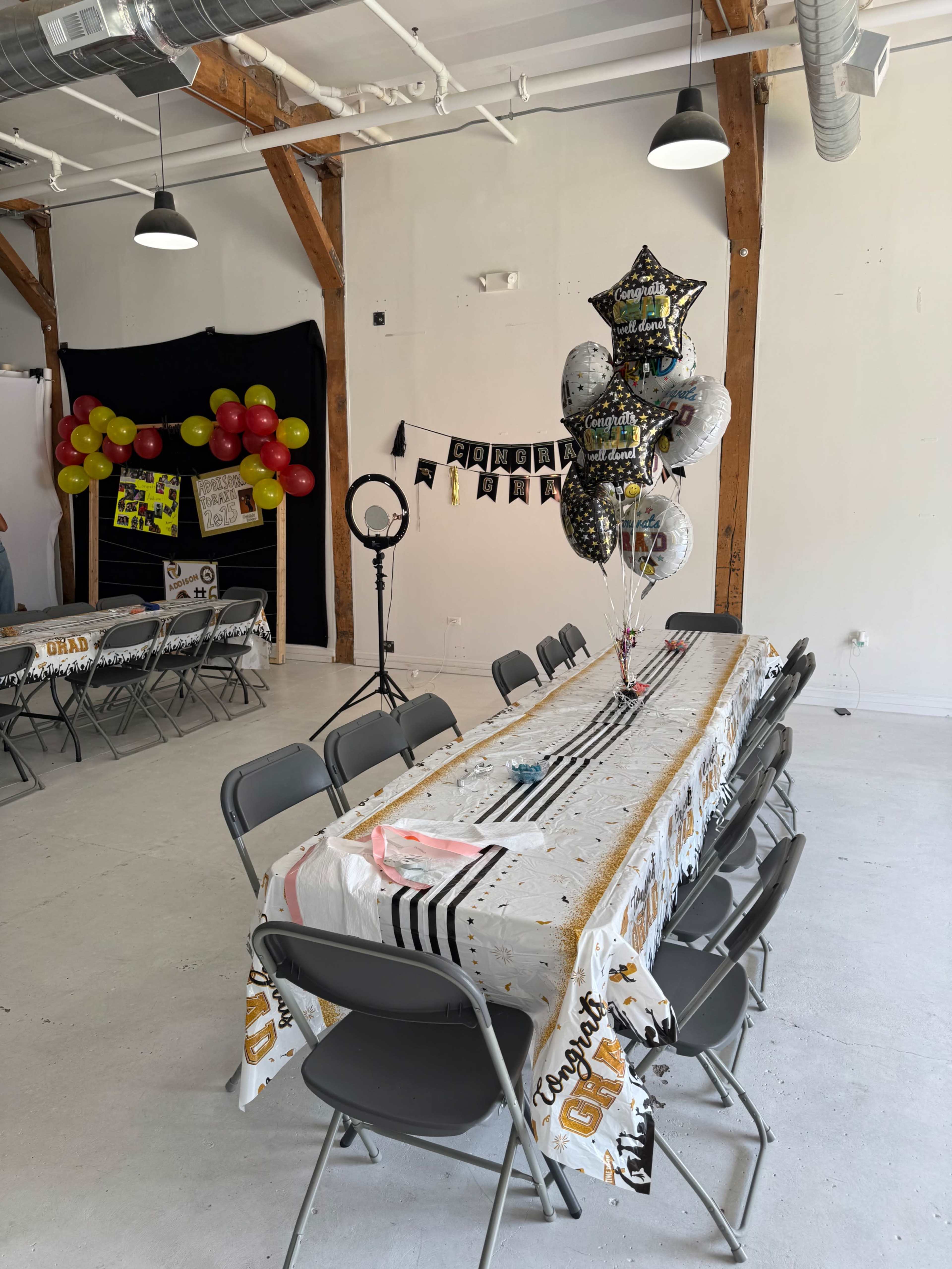 A long table with a celebratory tablecloth is set up in a room with wooden beams, surrounded by folding chairs and decorated with balloons and a banner.