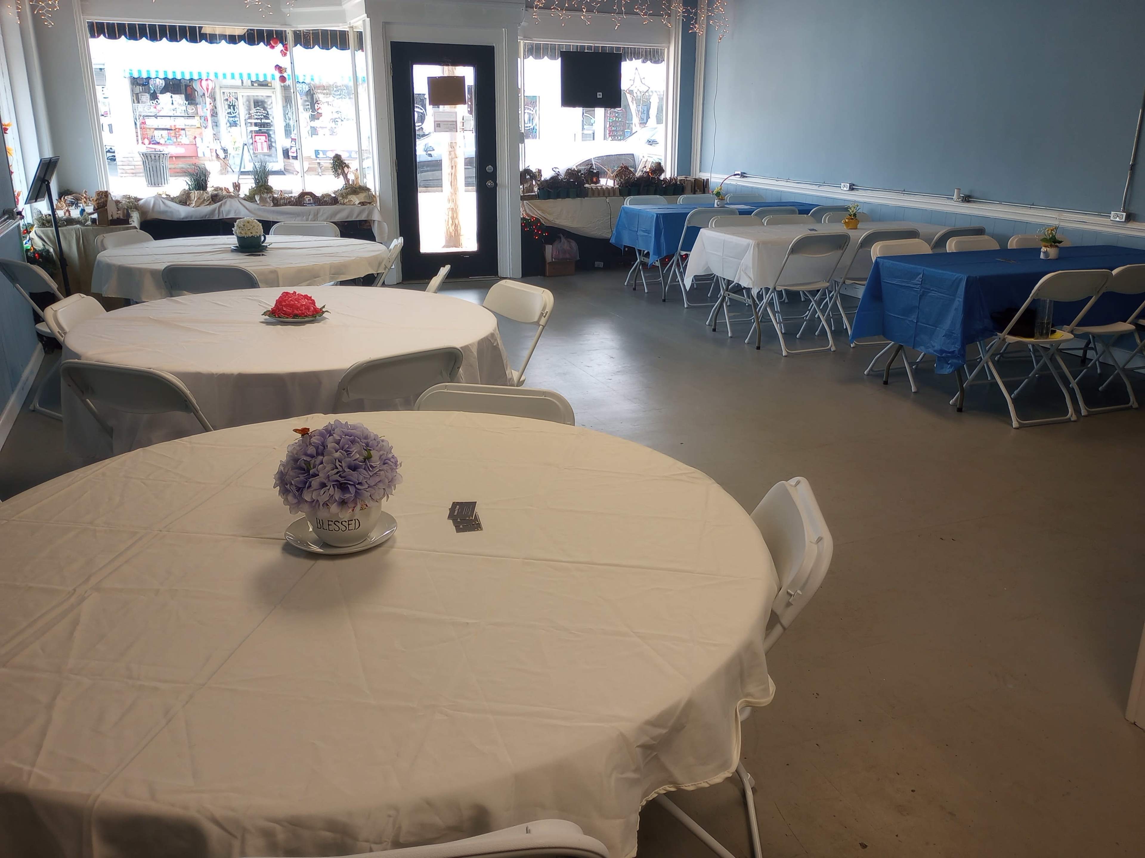 The image shows a spacious indoor area with several round tables covered in white and blue tablecloths, arranged for an event, and a large window displaying an outdoor scene.