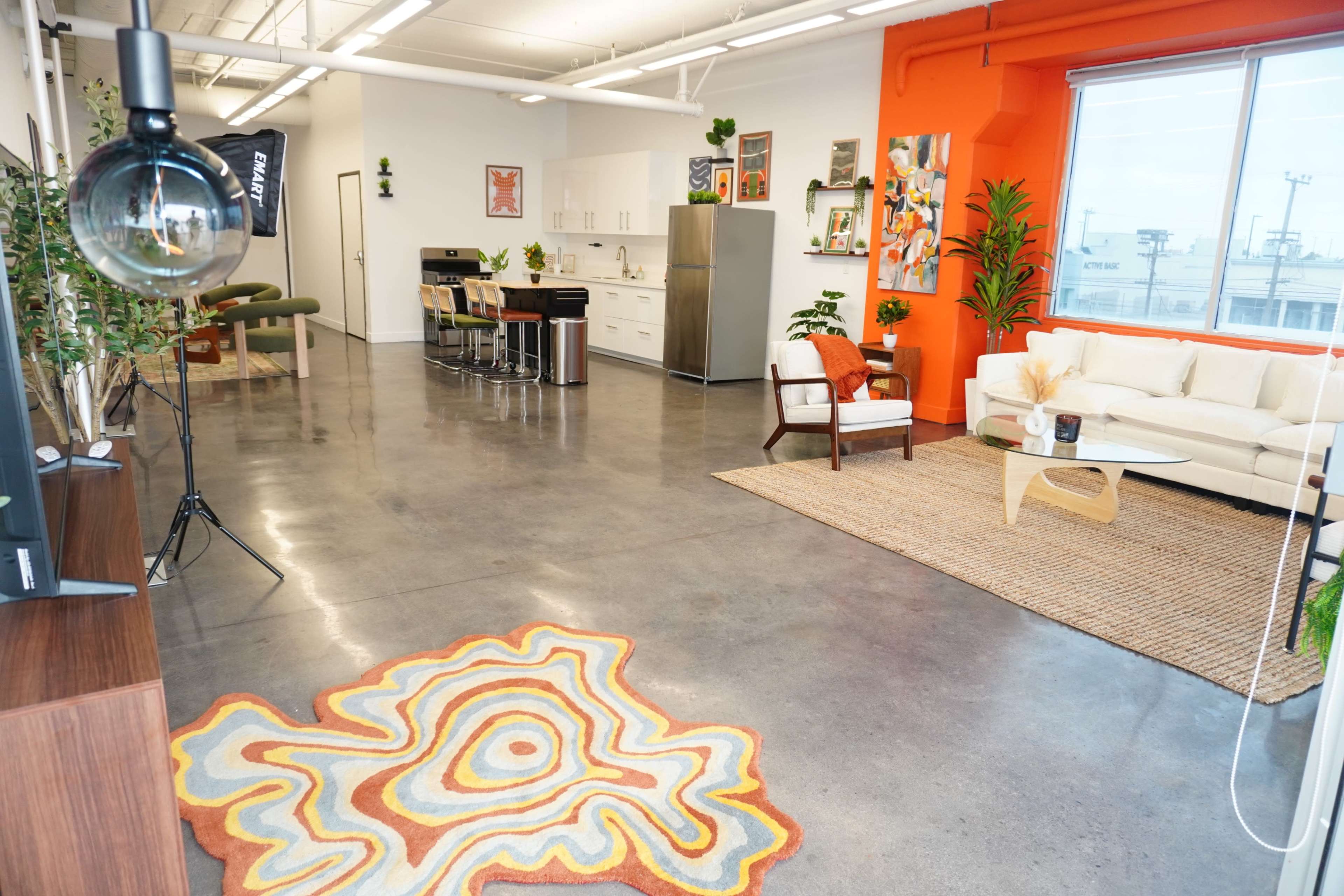 The image shows a modern, open-concept interior featuring a kitchen area, seating space with a white sofa, and decorative plants against an orange accent wall.