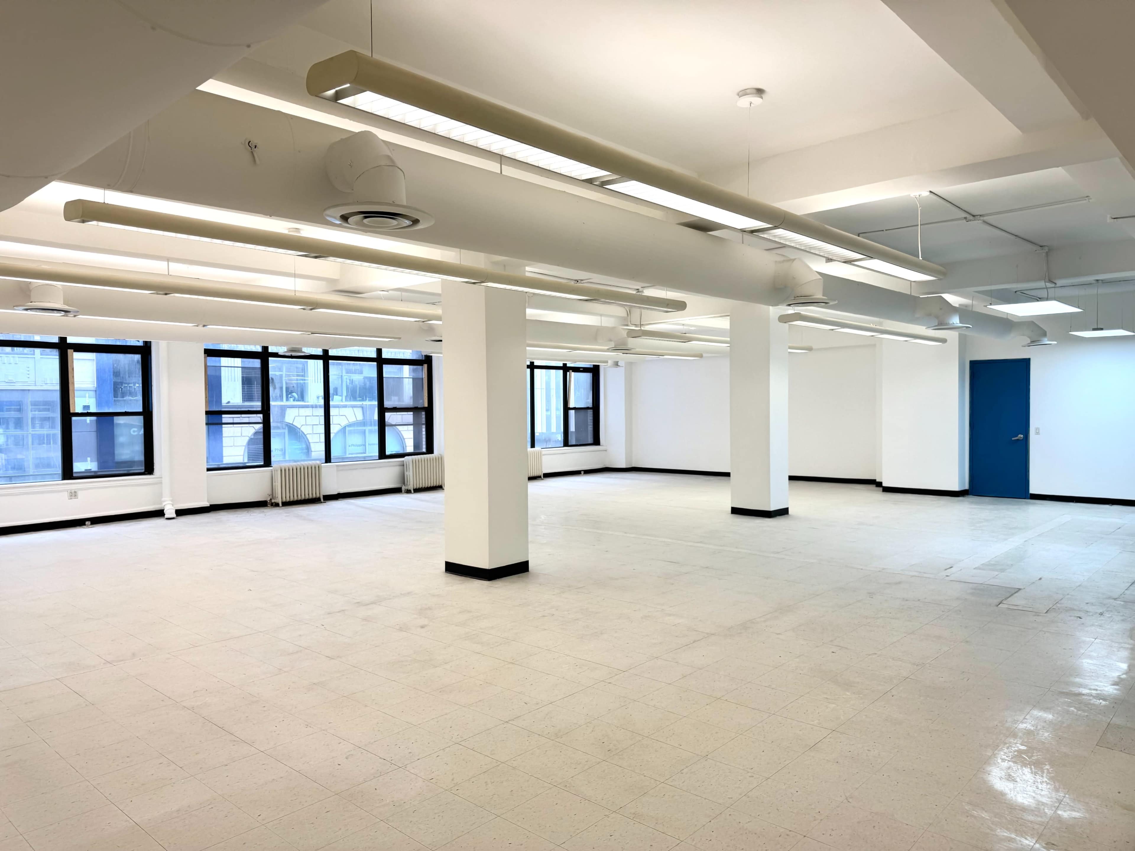 The image shows a large, empty room with white walls, several fluorescent lights, and multiple windows allowing natural light.