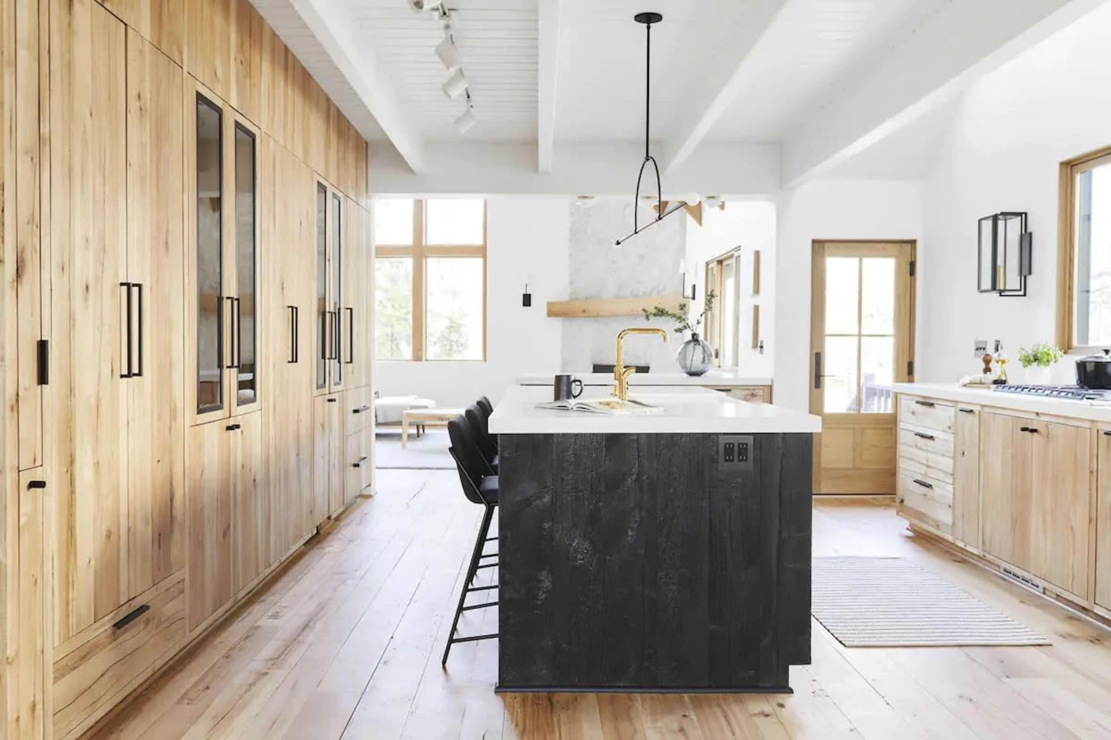 A modern kitchen features wooden cabinetry, a large island with a dark finish, and ample natural light from large windows.