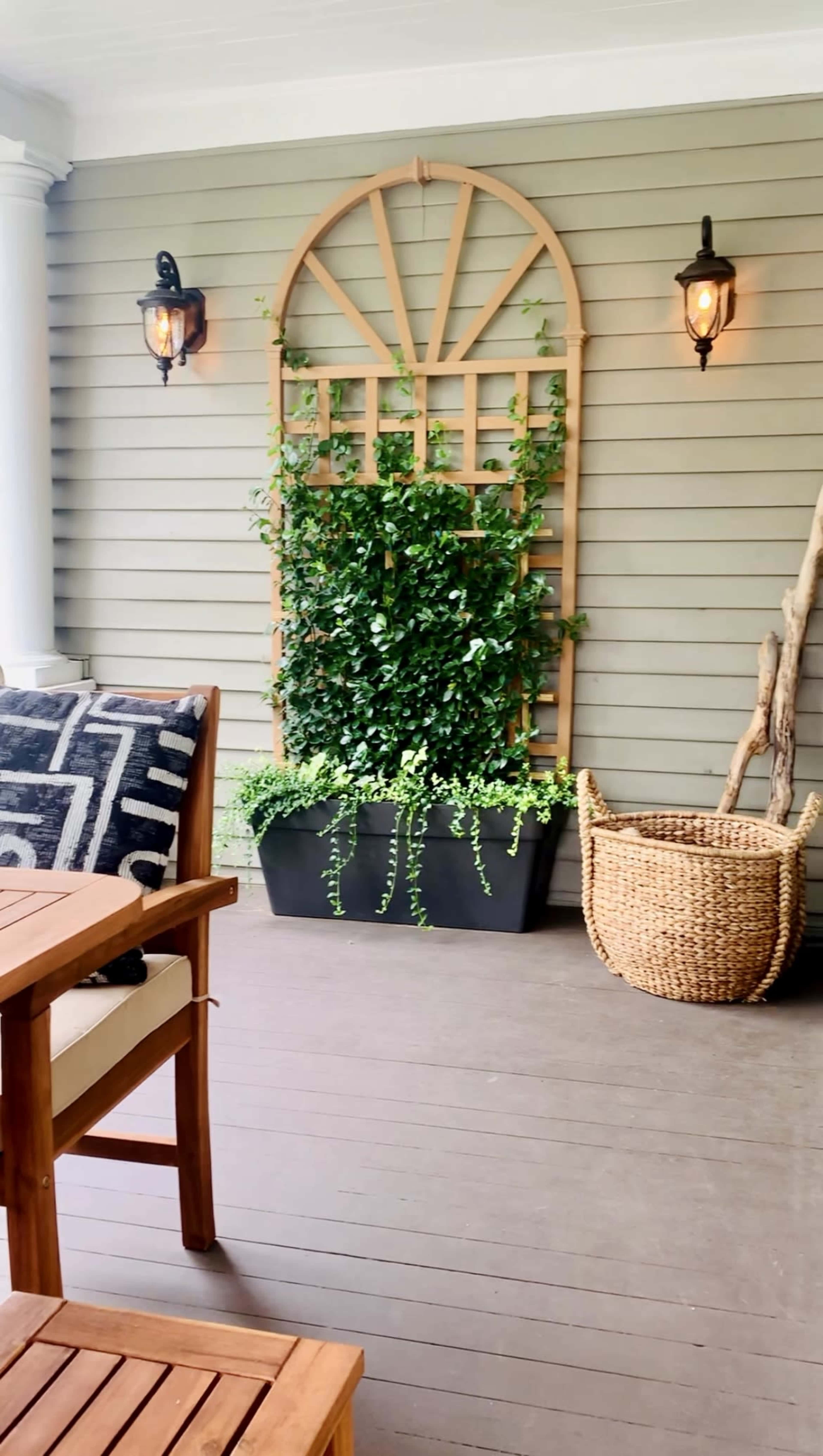 The image shows a porch area featuring a wooden table and chairs, a trellis with greenery, a wicker basket, and wall-mounted lanterns against a gray siding background.