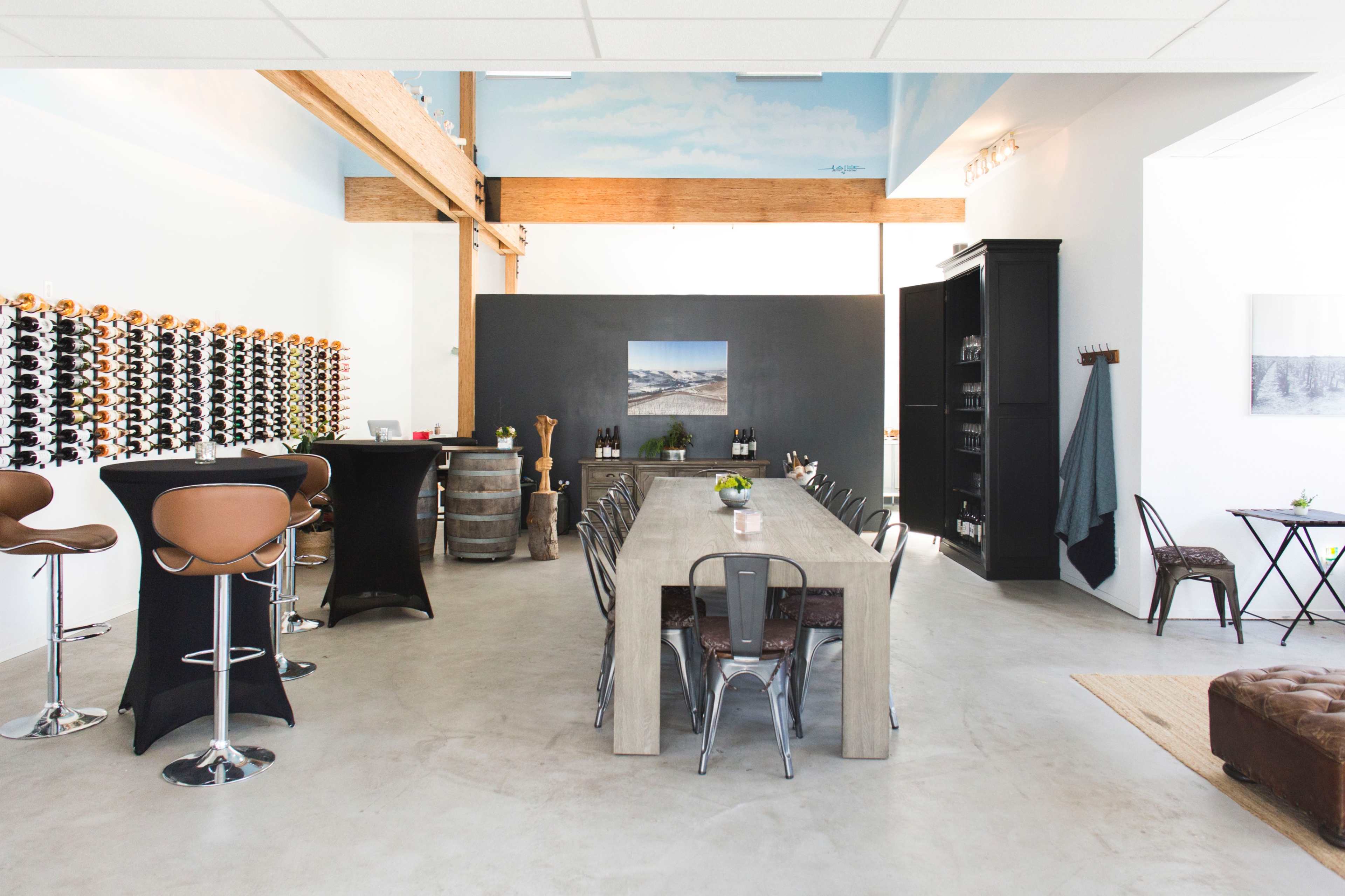 A modern wine tasting room featuring a long wooden table, bar stools, a wall of wine bottles, and minimalist decor.