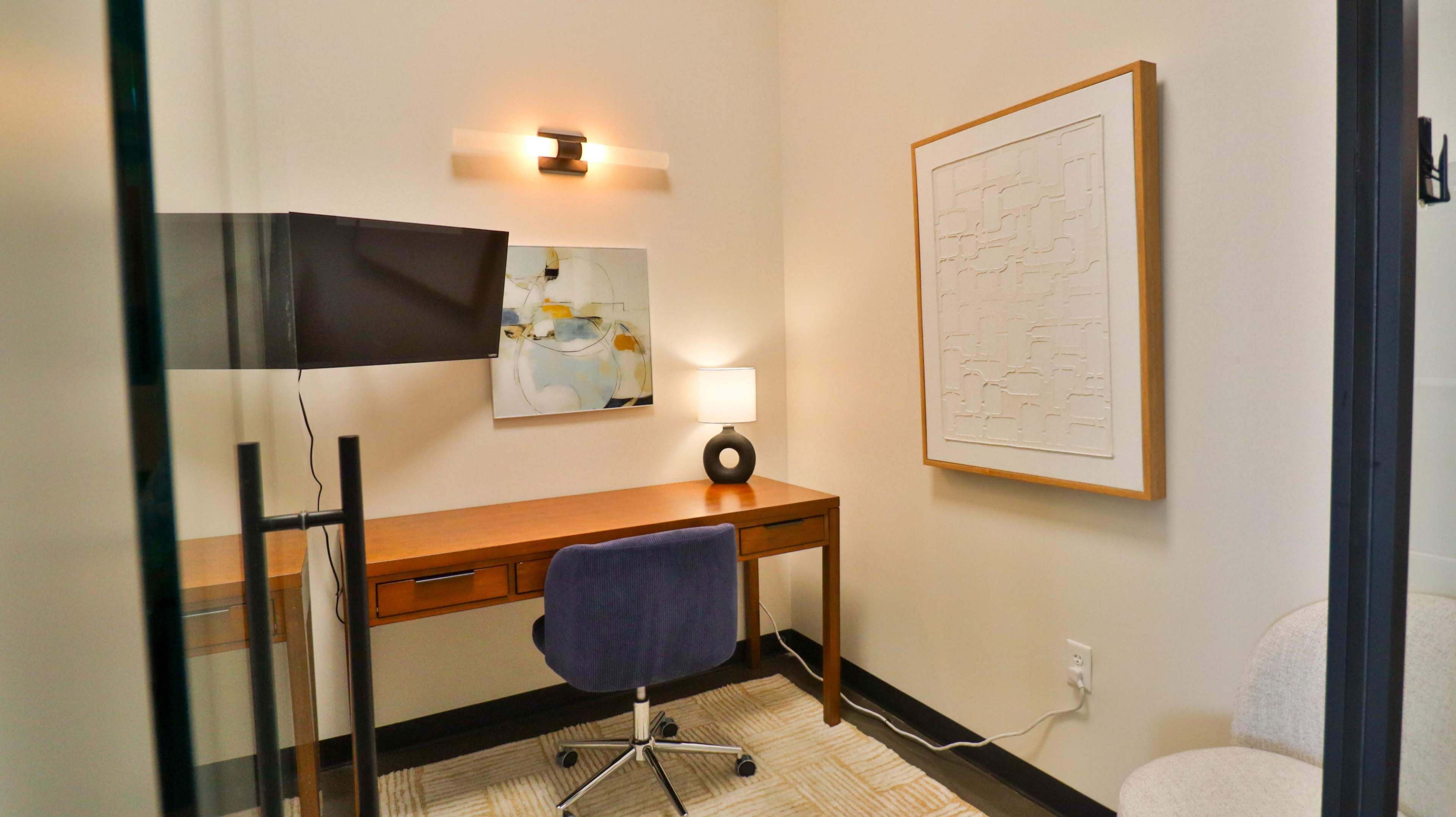 The image depicts a small office space with a desk, a chair, a television mounted on the wall, and framed artwork.