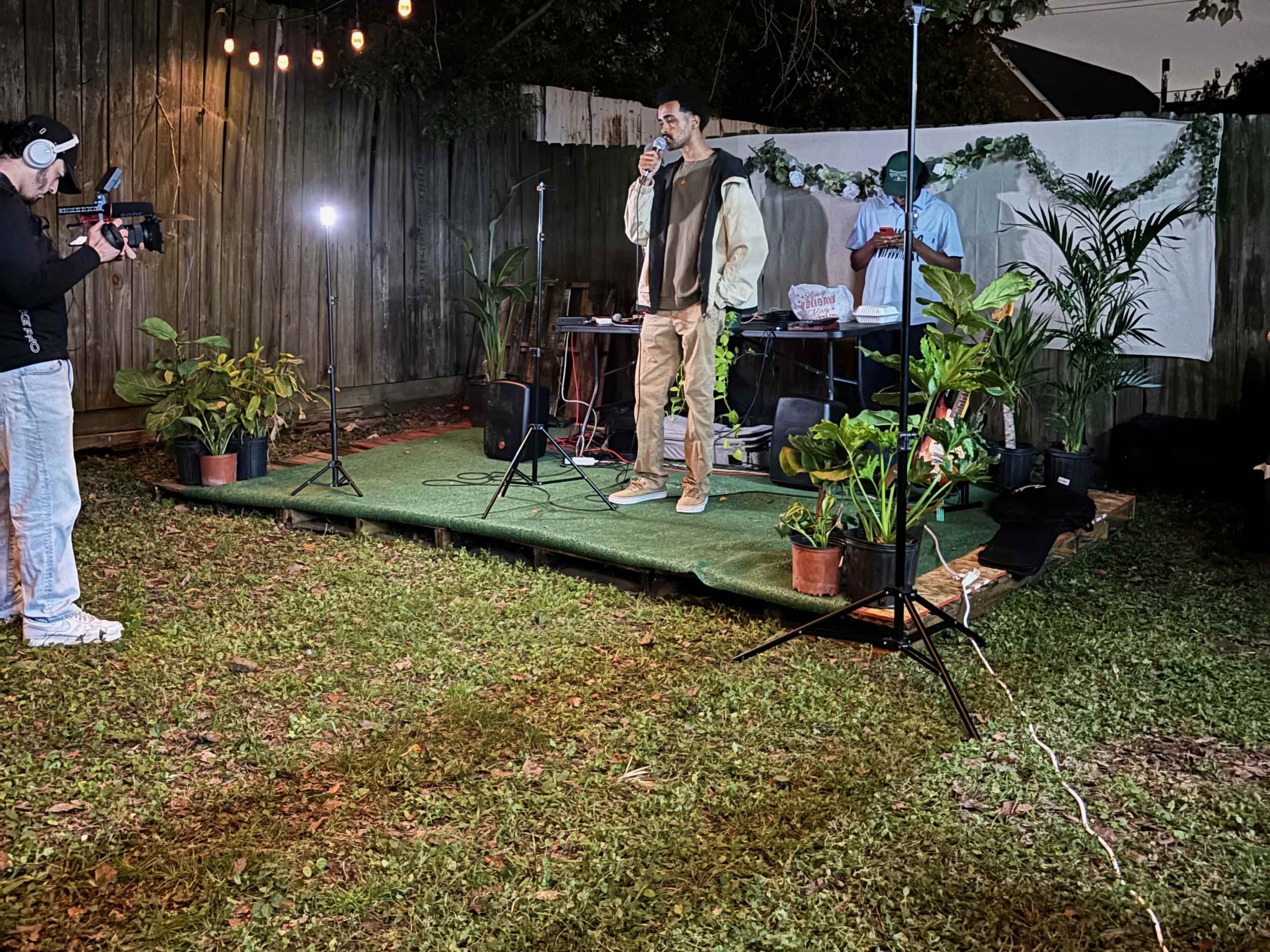 A man stands on a small stage in a backyard, speaking into a microphone while another person records him with a camera.