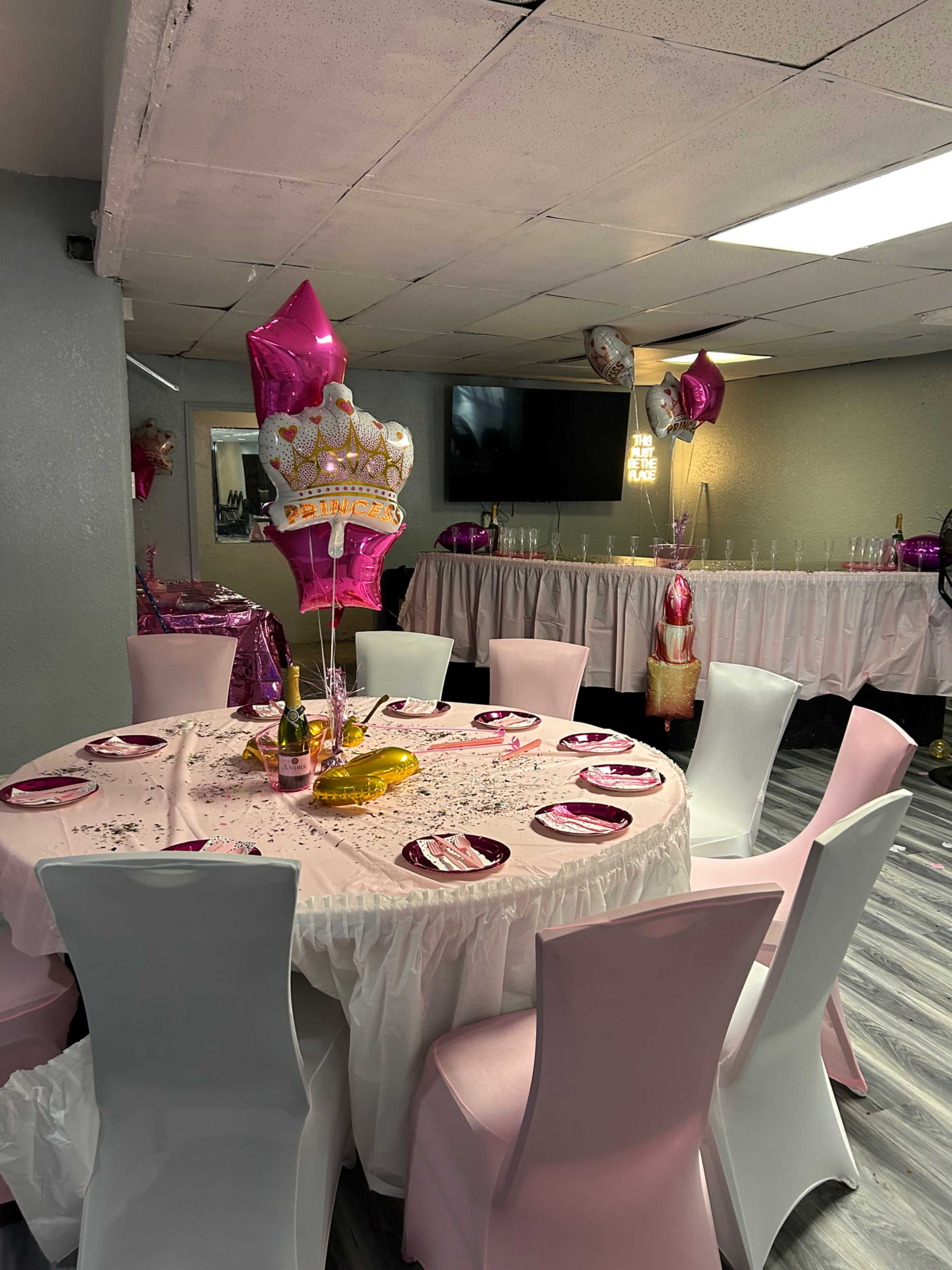 A round table set with pink and gold decorations, surrounded by white chairs, is prepared for a celebration in a decorated room.