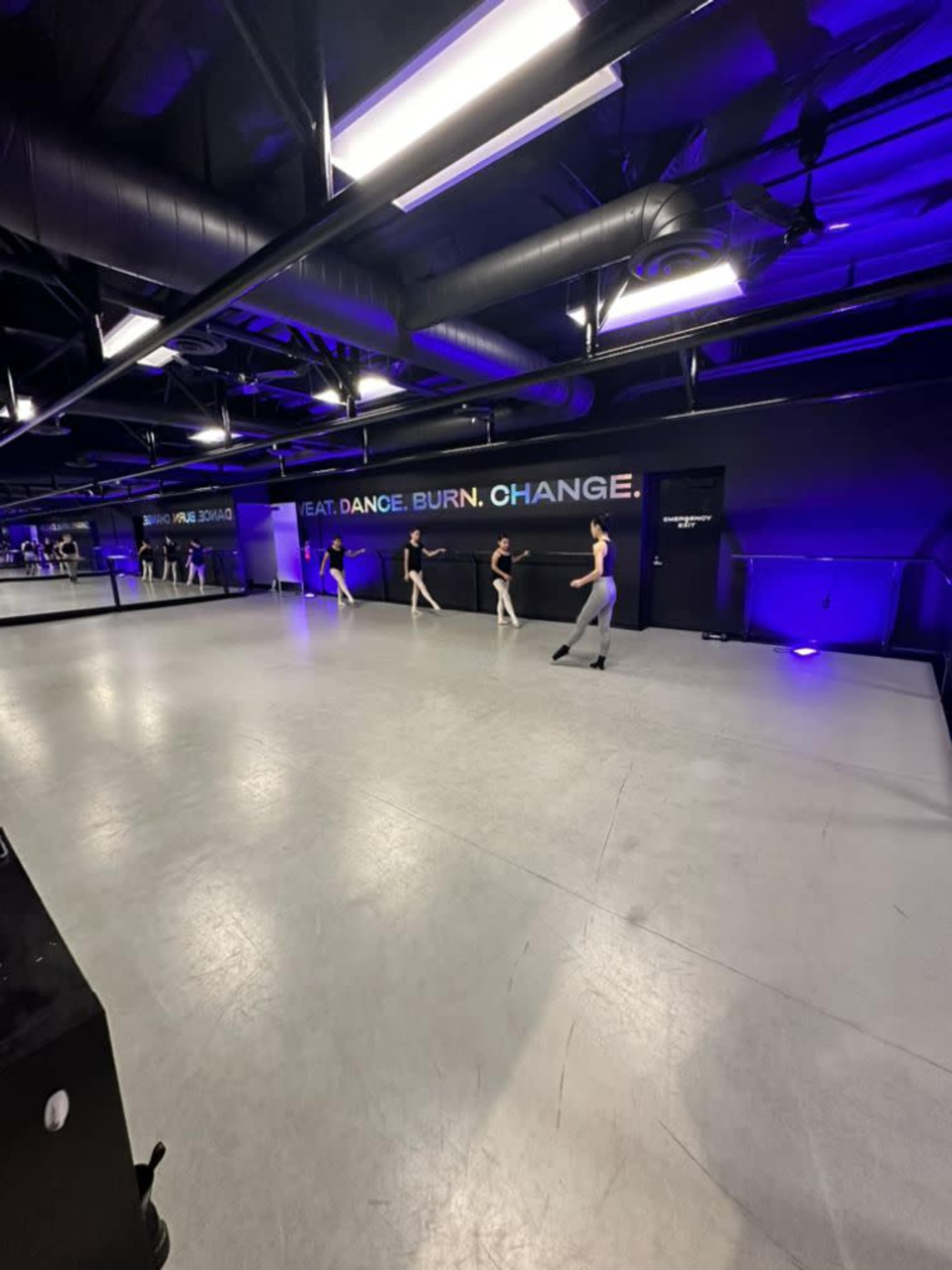 A group of dancers practice in a studio with mirrors and a black wall displaying the words "FEAT. DANCE. BURN. CHANGE."