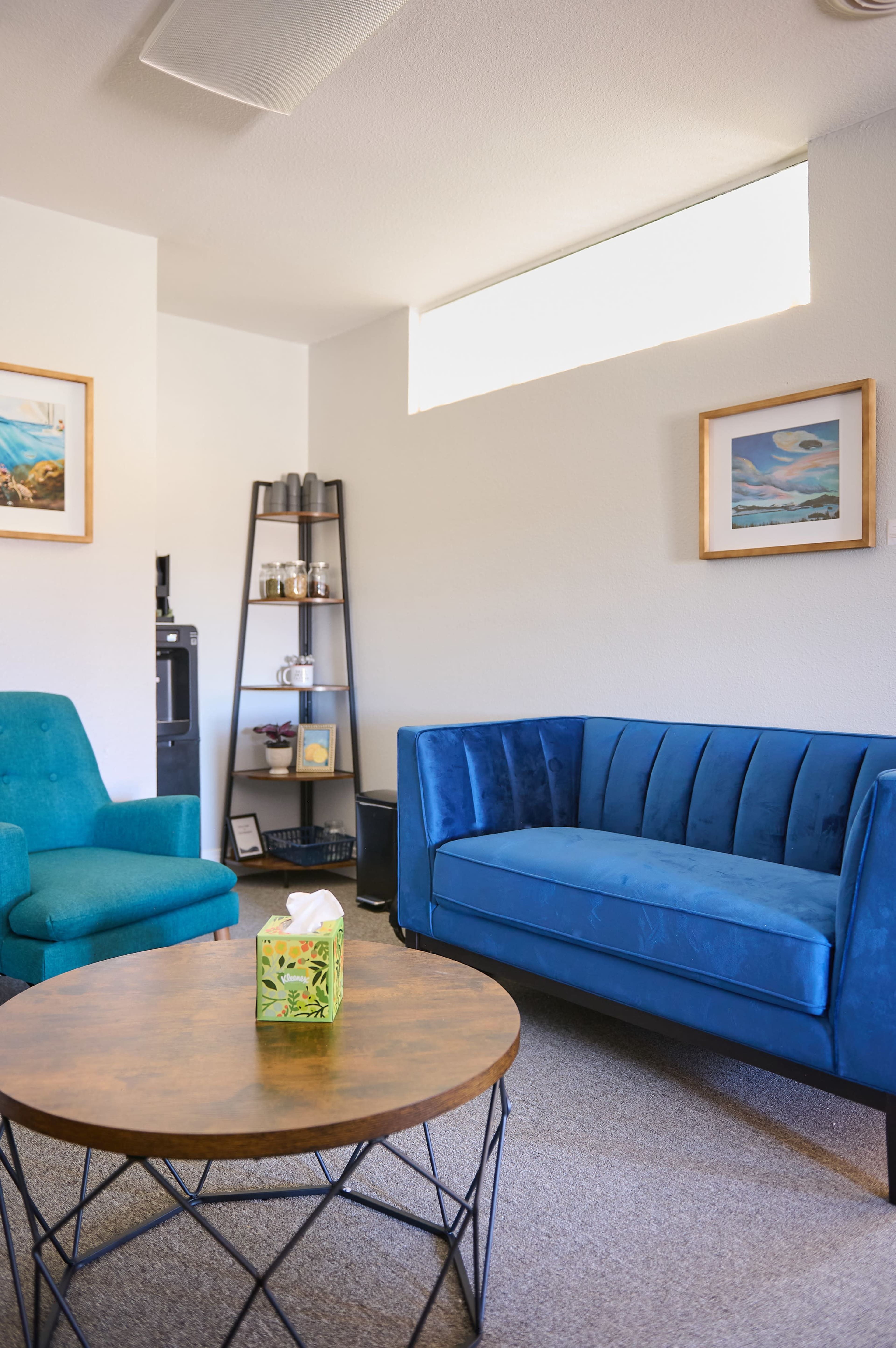 The image shows a modern living room with a blue couch, a teal armchair, a round wooden table, and decorative wall art.