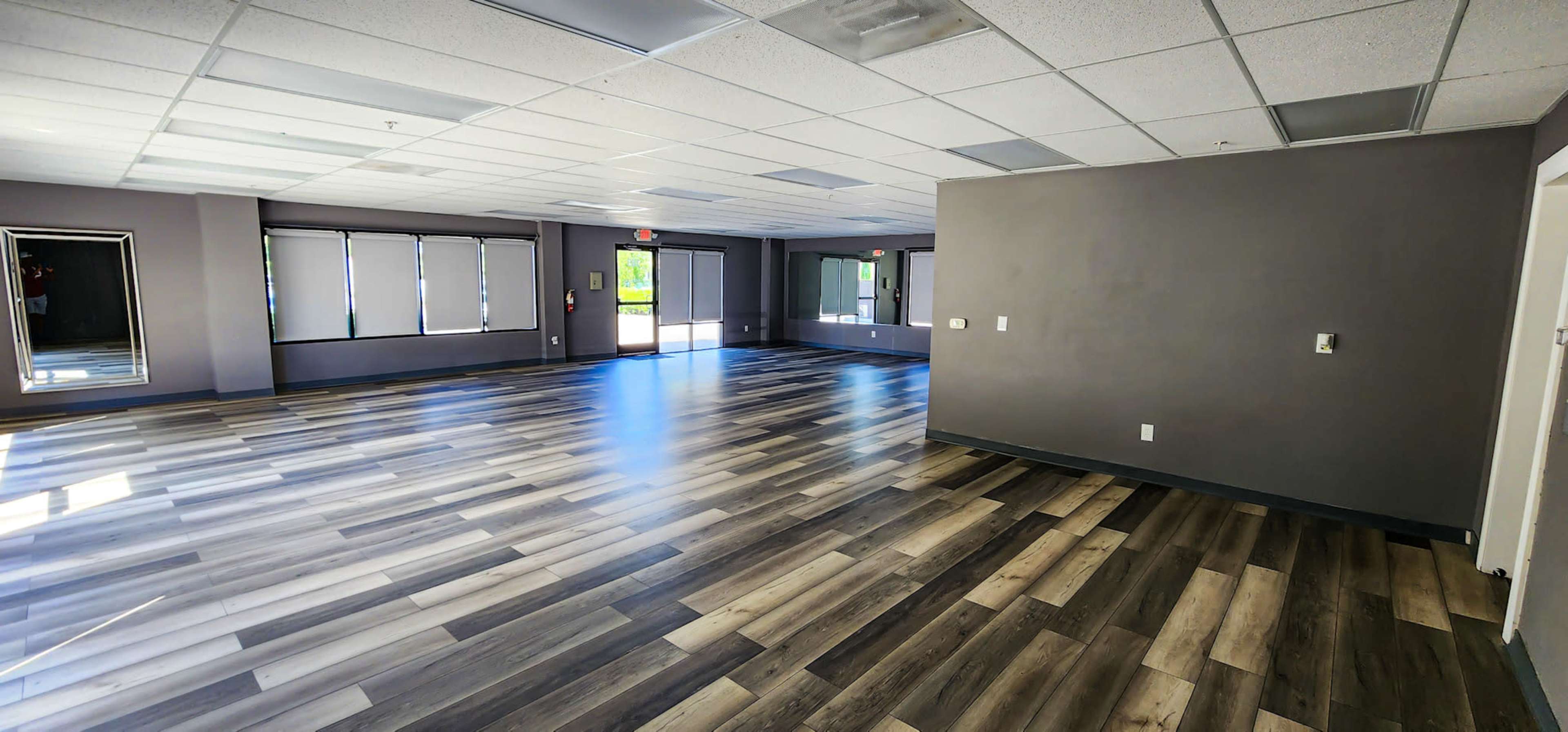 A spacious, well-lit room with large windows and a floor featuring alternating light and dark wooden planks.