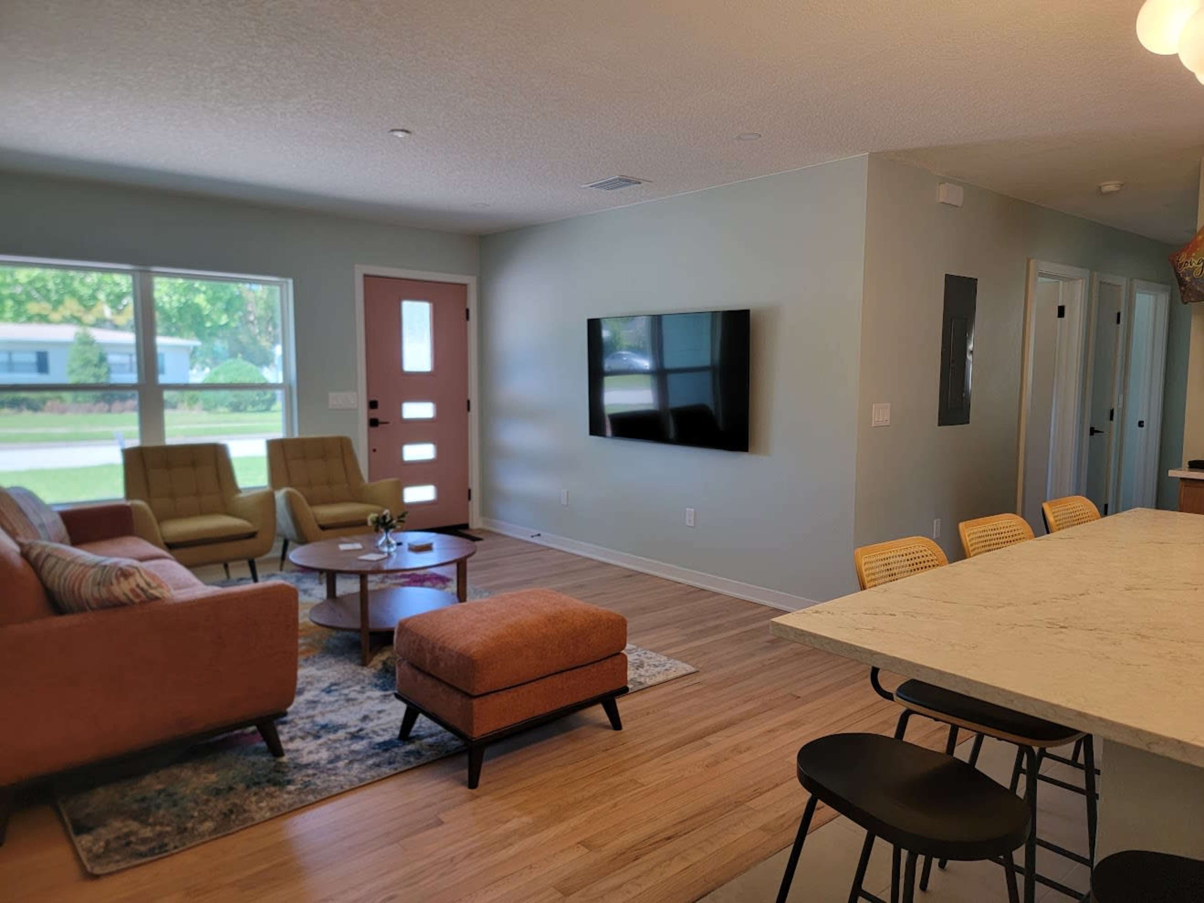 The image shows a modern living room with a couch, chairs, a coffee table, and a wall-mounted television, adjacent to a kitchen area with bar stools.