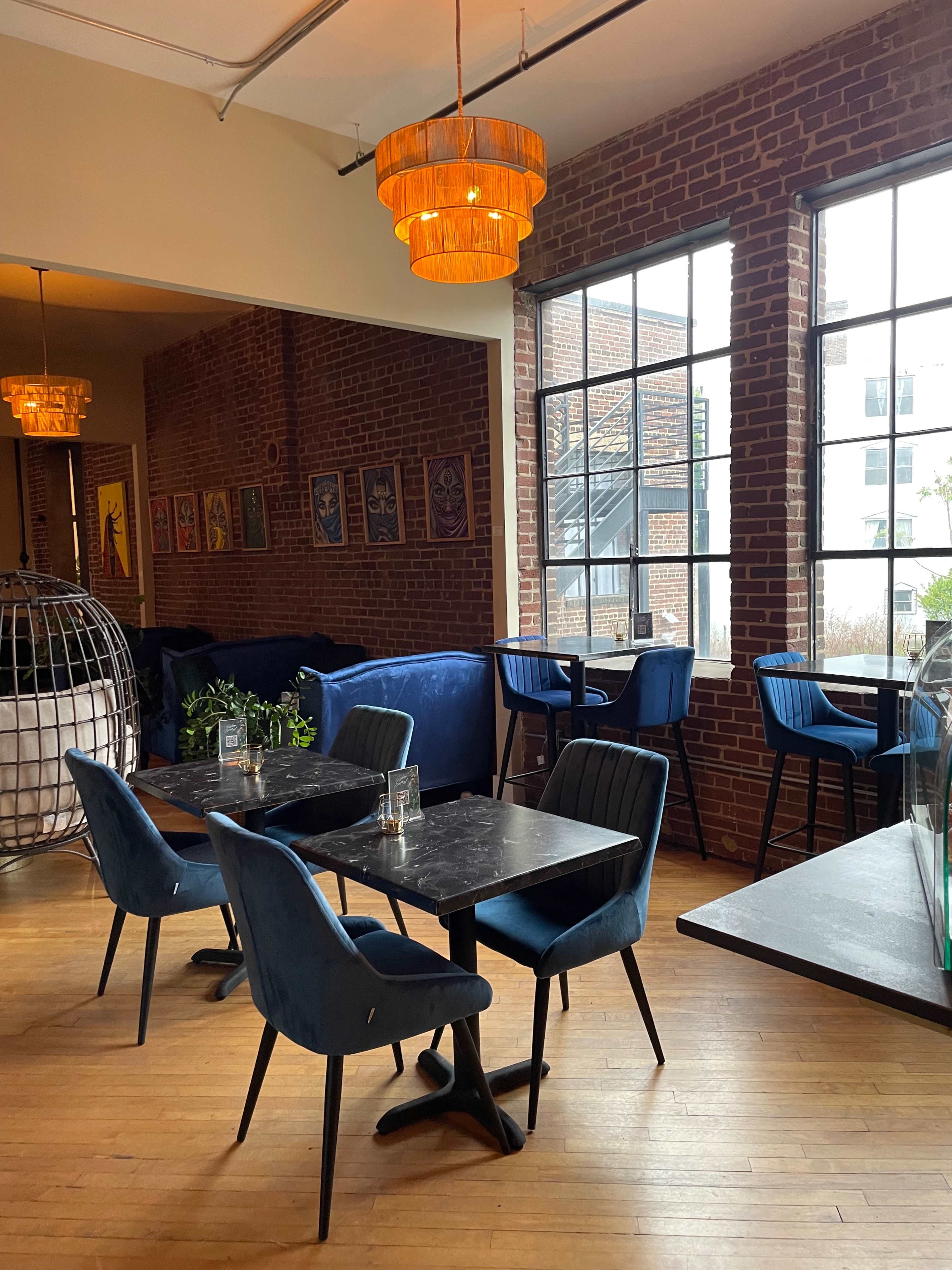 A modern cafe interior with brick walls, blue upholstered chairs, a large round pendant light, and tables set for dining.