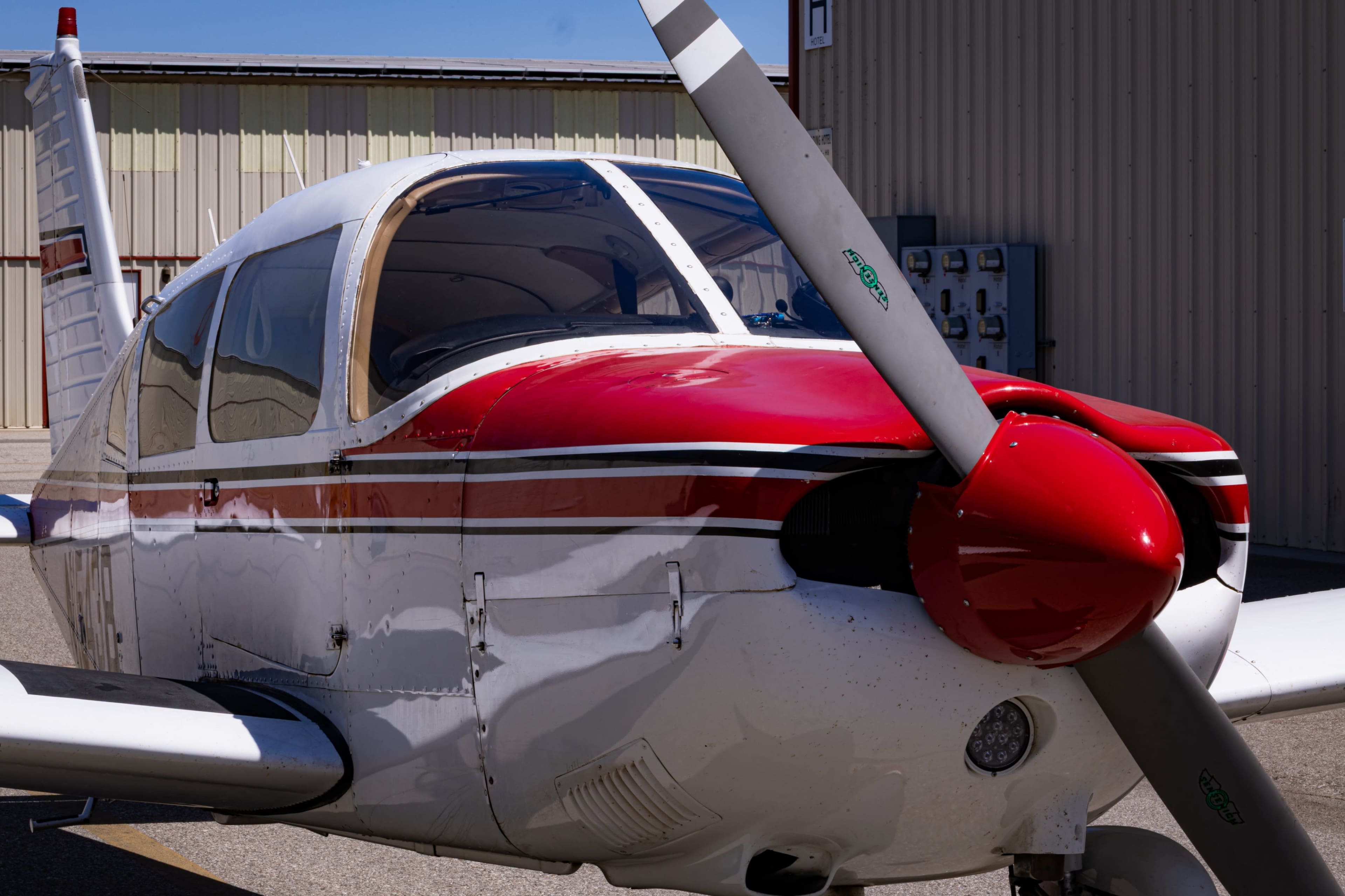 A red and white single-engine aircraft is parked on the tarmac beside a hangar.