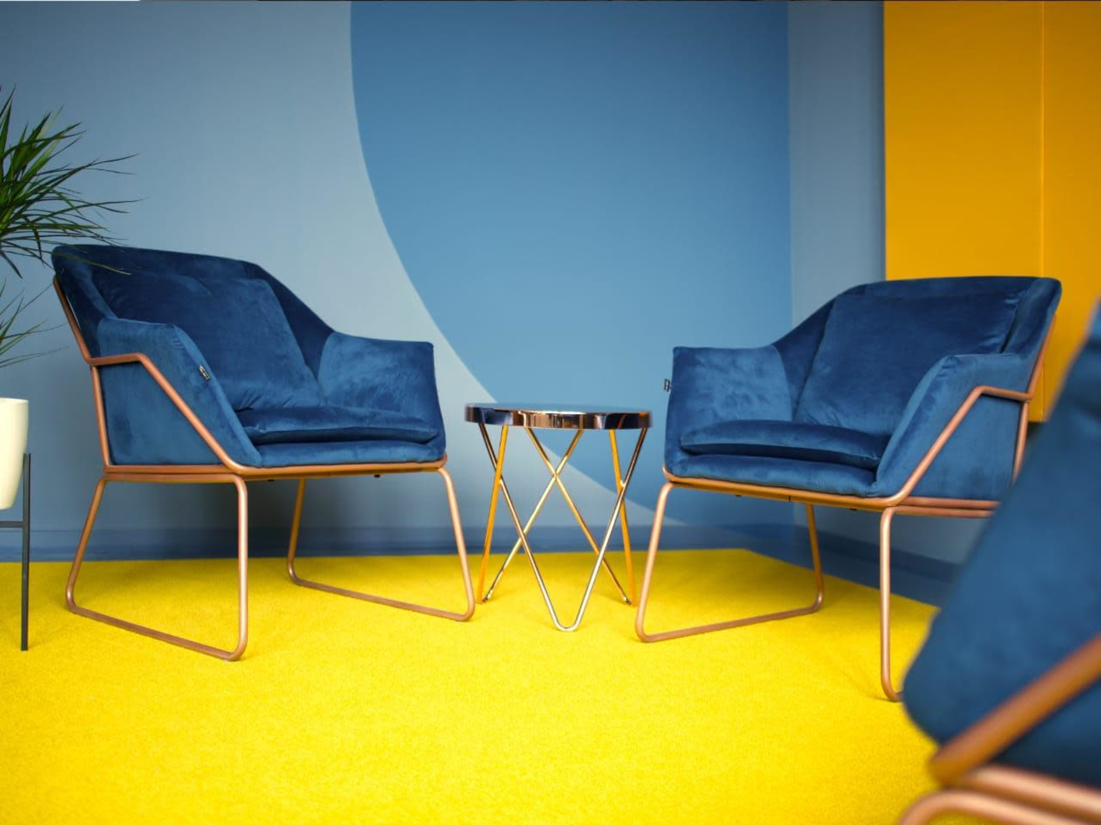 Two blue velvet chairs with metallic frames are positioned opposite each other next to a small, round table on a bright yellow carpet in a room with blue and yellow walls.