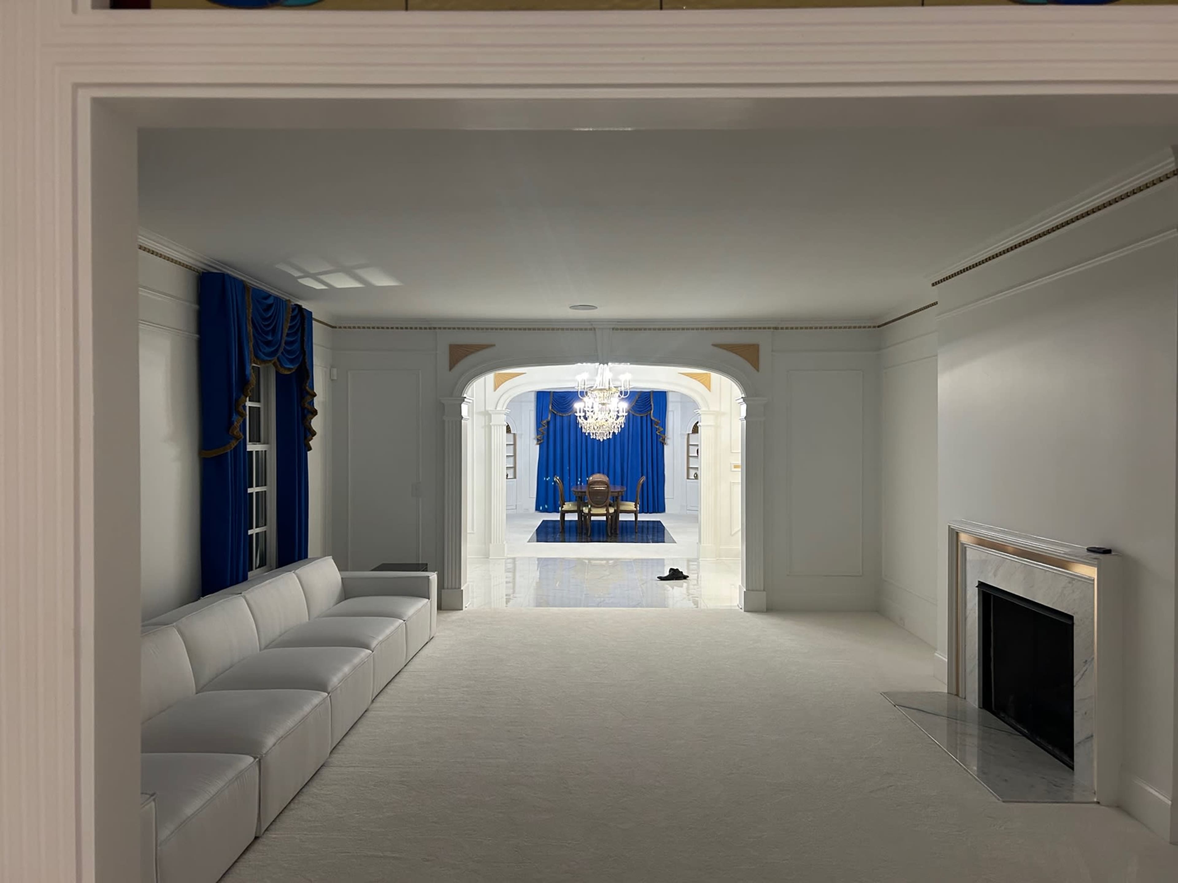The image shows a white room leading to another space with blue curtains and a chandelier, featuring a seating area with a white couch and a black shoe on the floor.