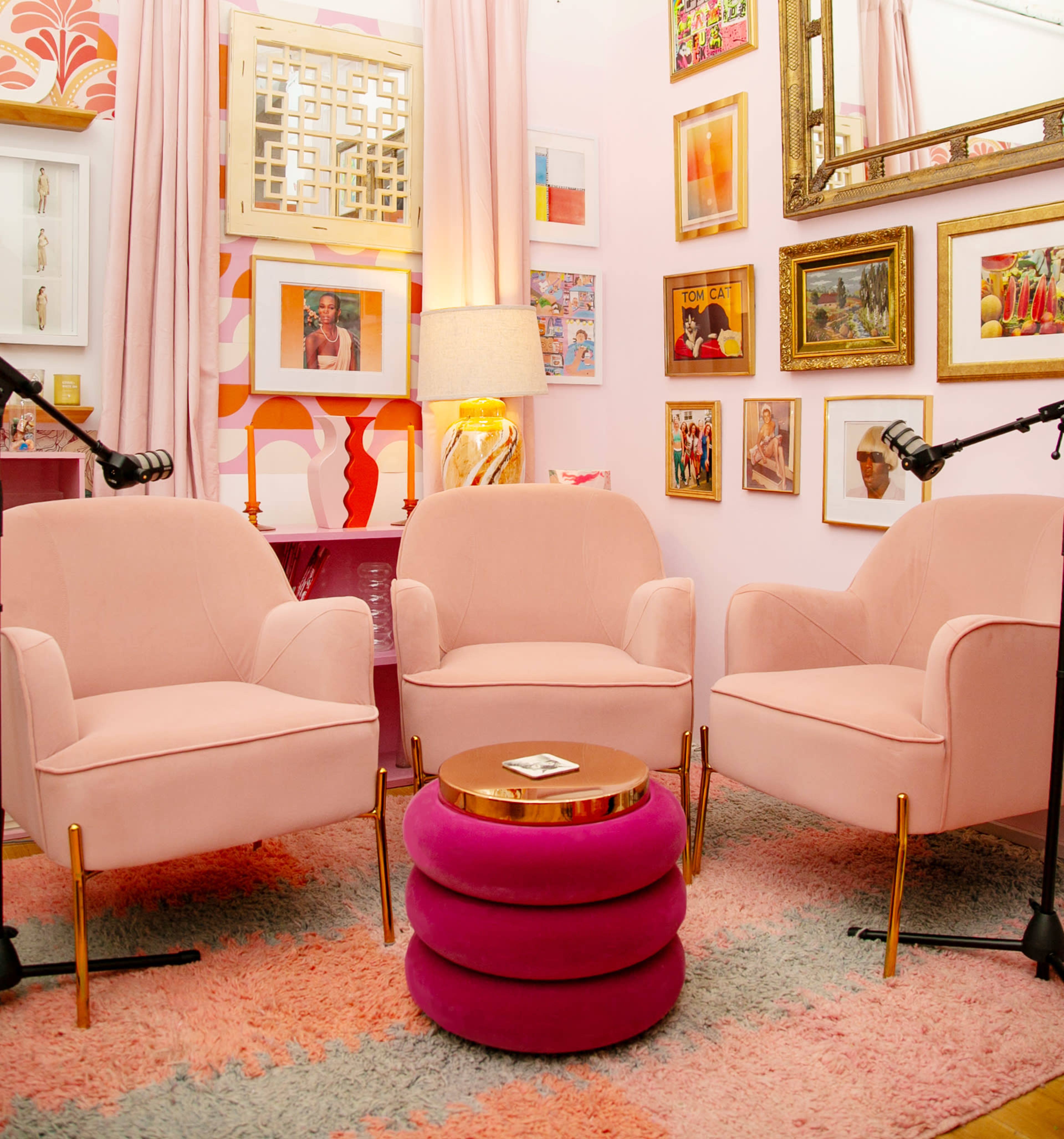 The image features a cozy room with two pink armchairs and a round, stacked pink table, surrounded by colorful framed artwork on the walls.
