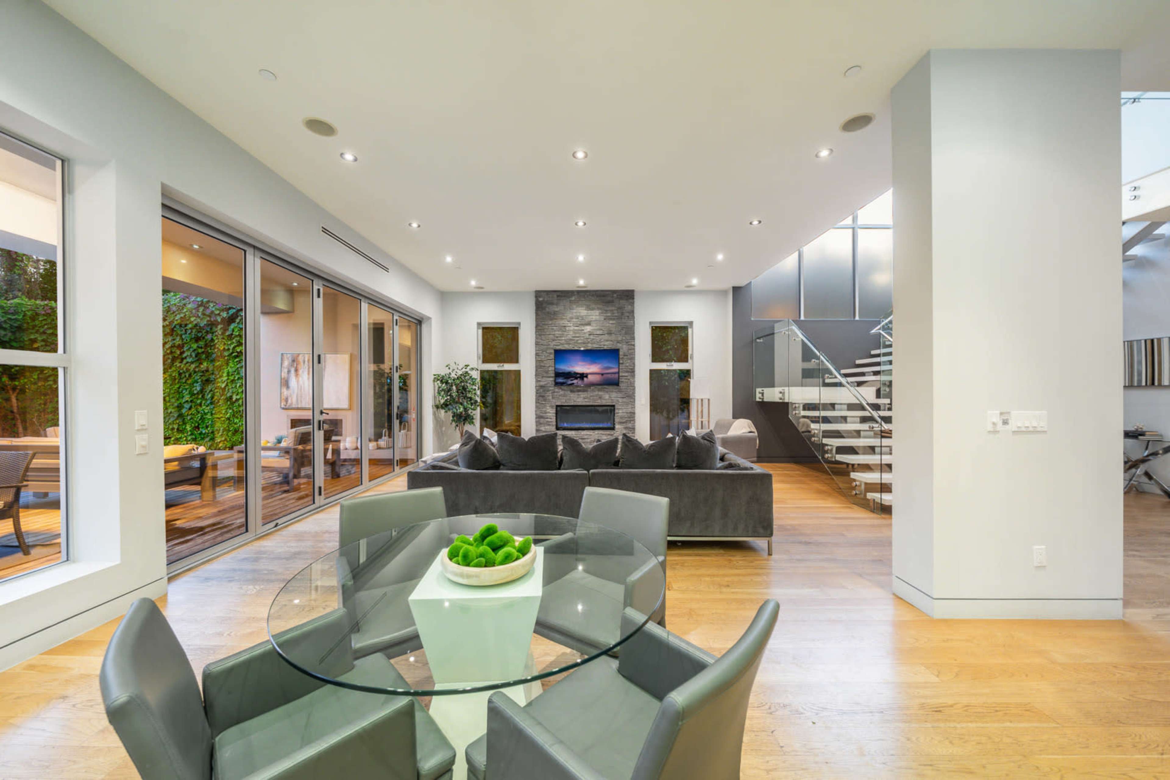 A modern living space features a glass dining table with a white centerpiece, a gray sectional sofa, and large glass doors opening to an outdoor area.