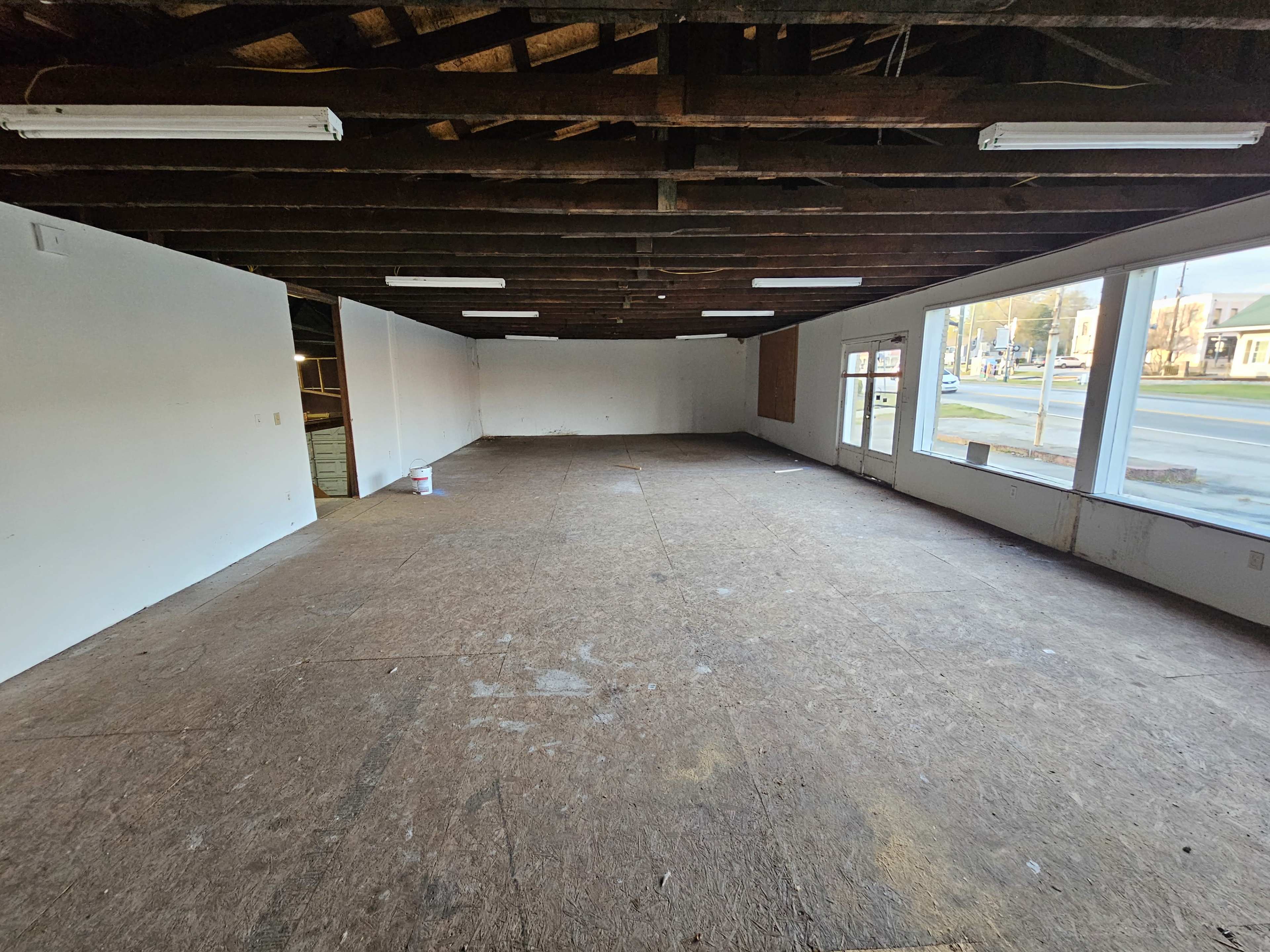 The image shows an empty commercial space with exposed wooden beams and a bare floor, featuring large front windows and a view of a street outside.