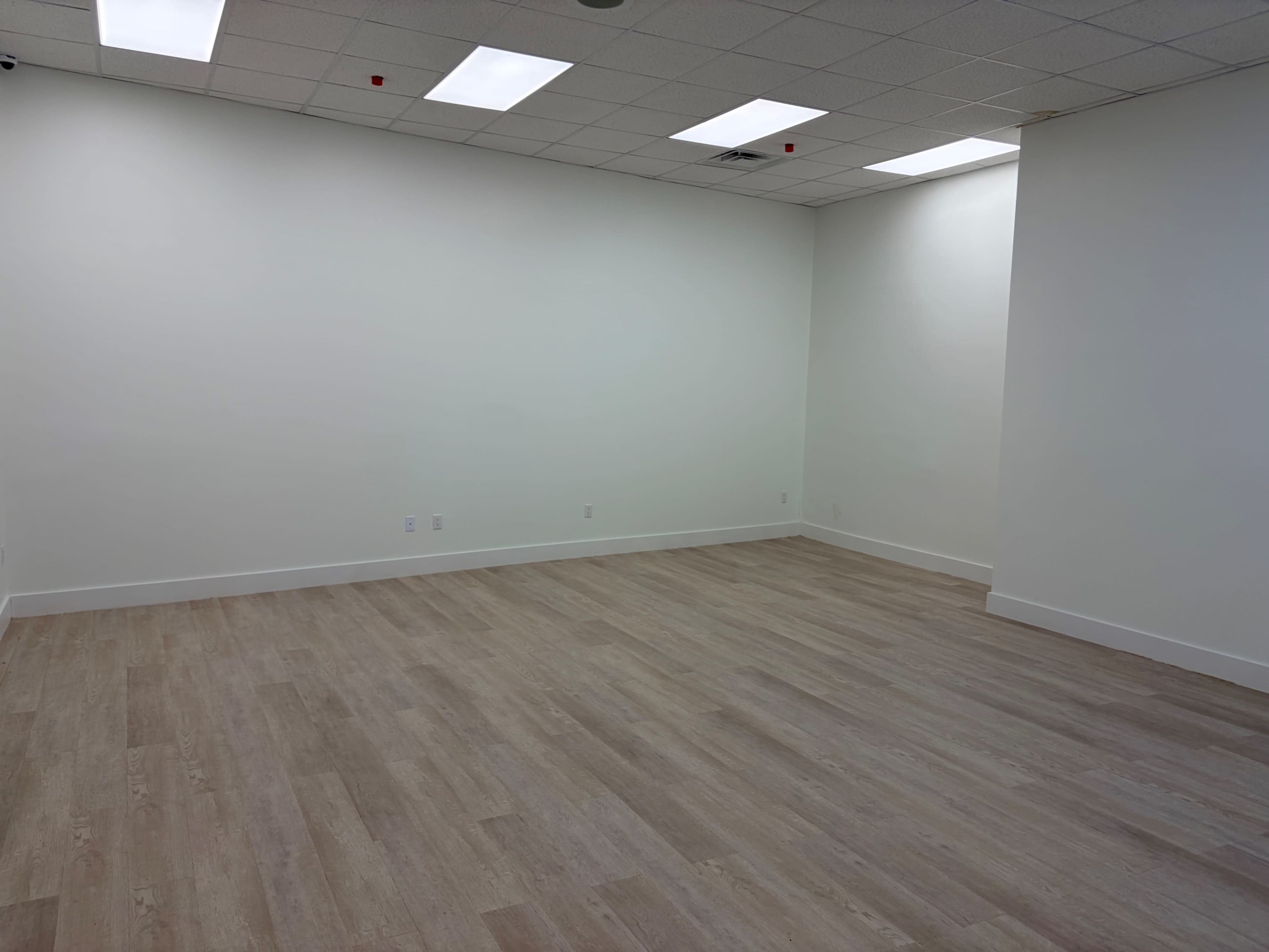 The image shows an empty room with light wooden flooring and white walls, illuminated by ceiling lights.