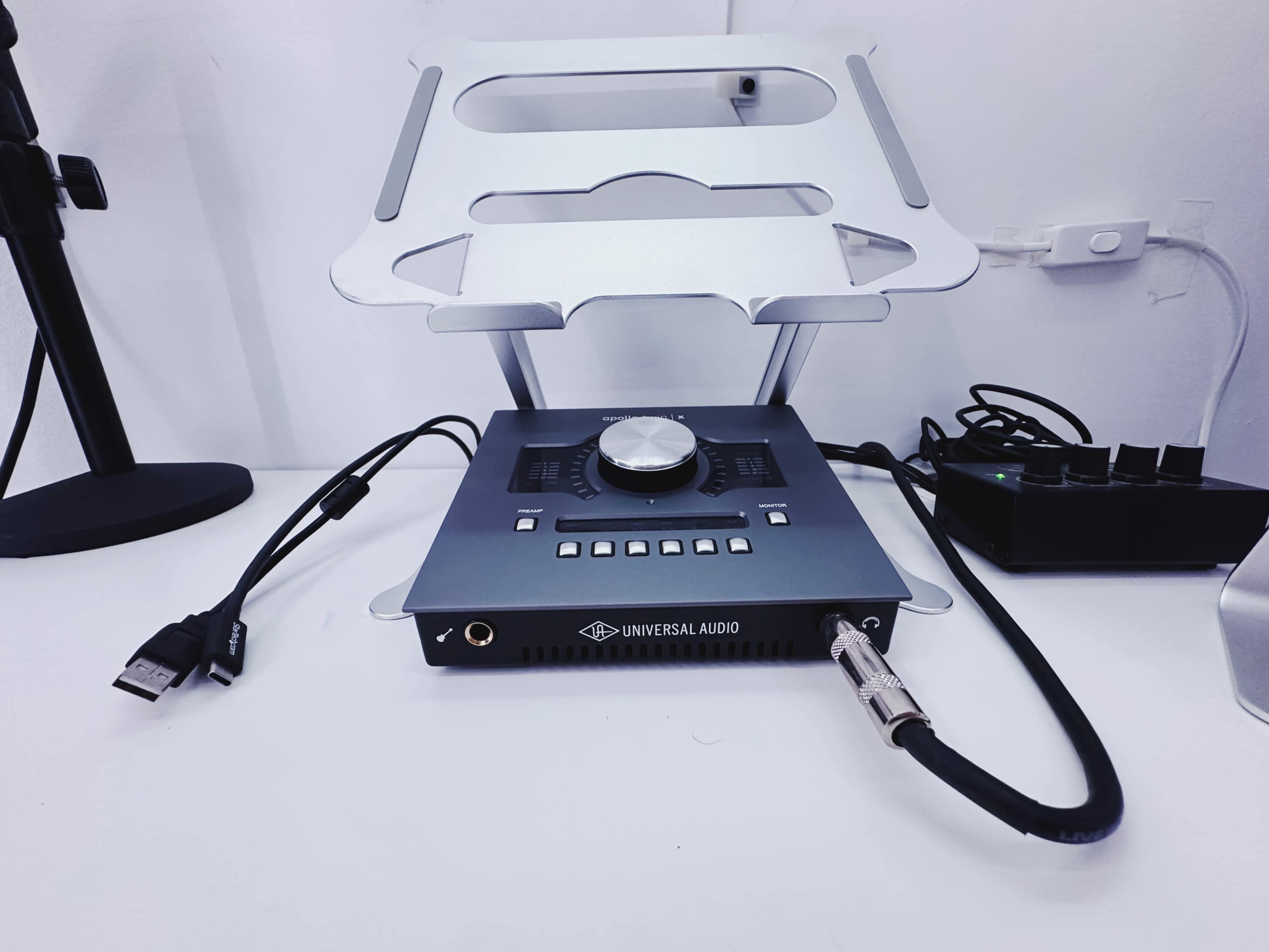 A Universal Audio audio interface sits on a silver stand among various cables and a power strip on a desk.