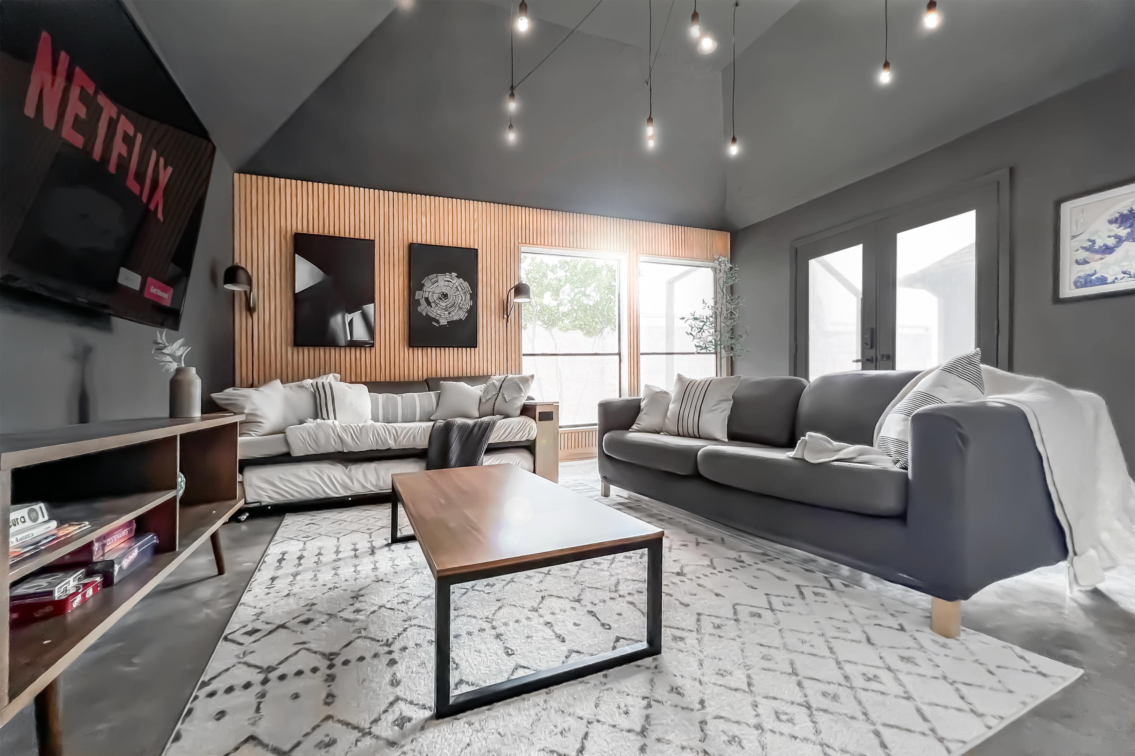 A modern living room features a gray sofa, patterned area rug, wooden accent wall, and a television displaying the Netflix logo.