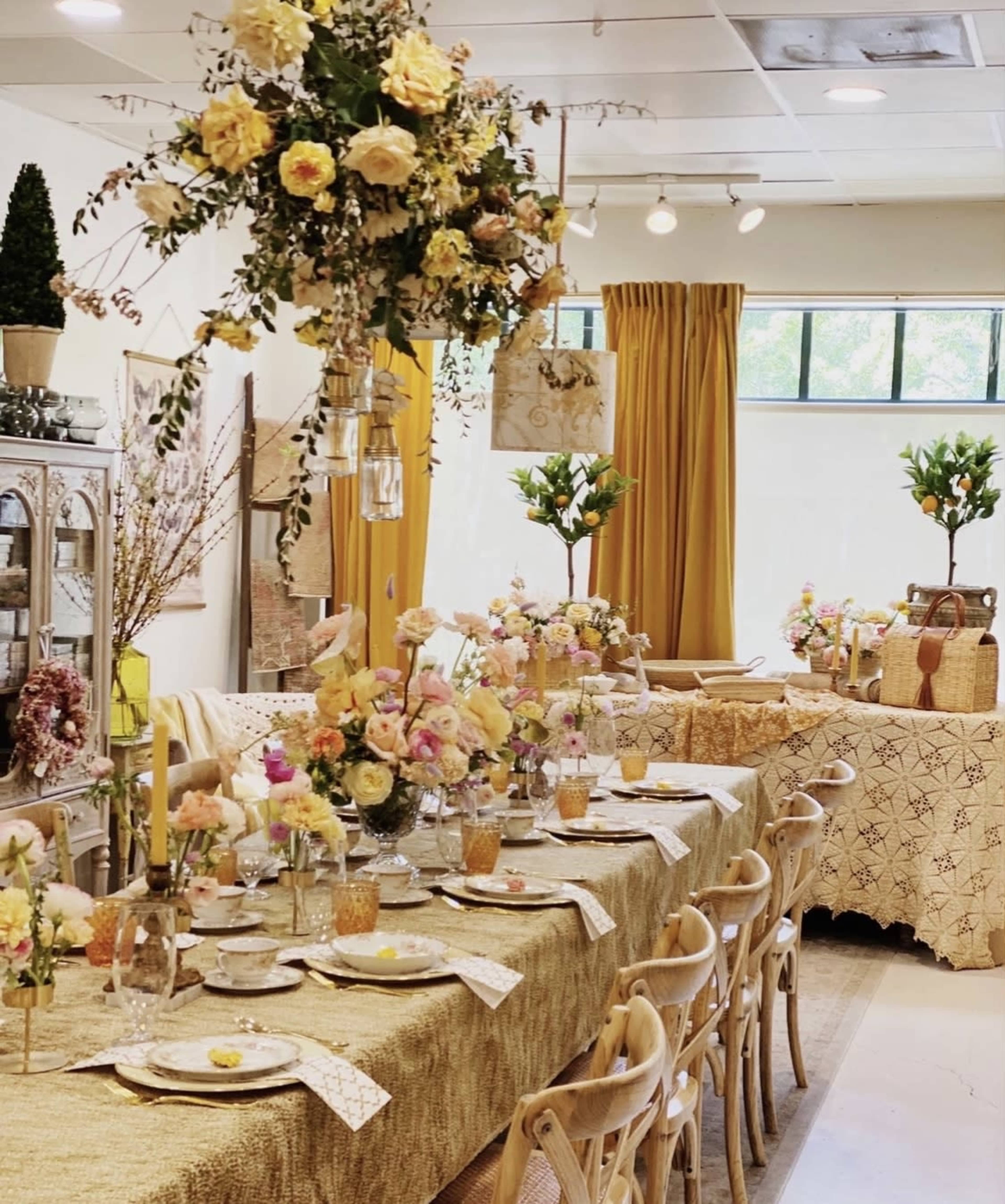 A decorated dining space featuring a long table set with floral centerpieces, glassware, and gold accents, surrounded by elegant chairs and adorned with additional floral arrangements and textiles.