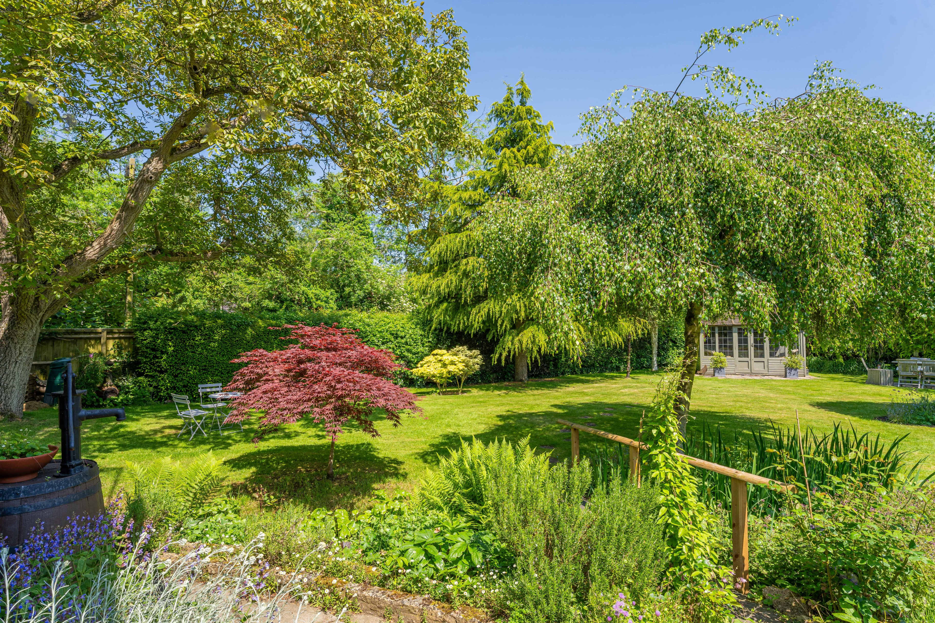 A well-maintained garden features a variety of trees, a Japanese maple, and a wooden gazebo in the background.