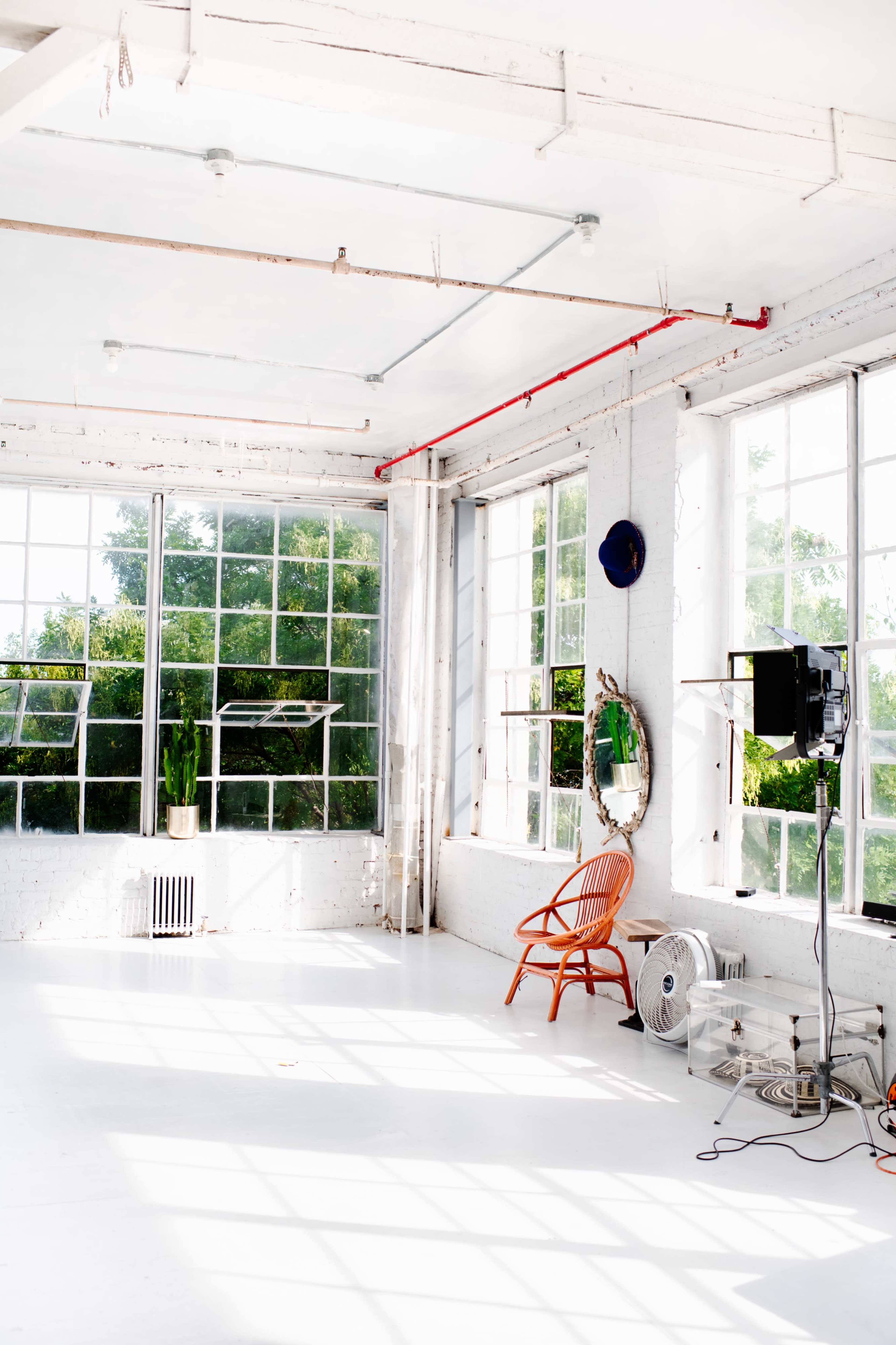 A spacious, well-lit studio with large windows, a single wooden chair, a fan, and a wall mirror.