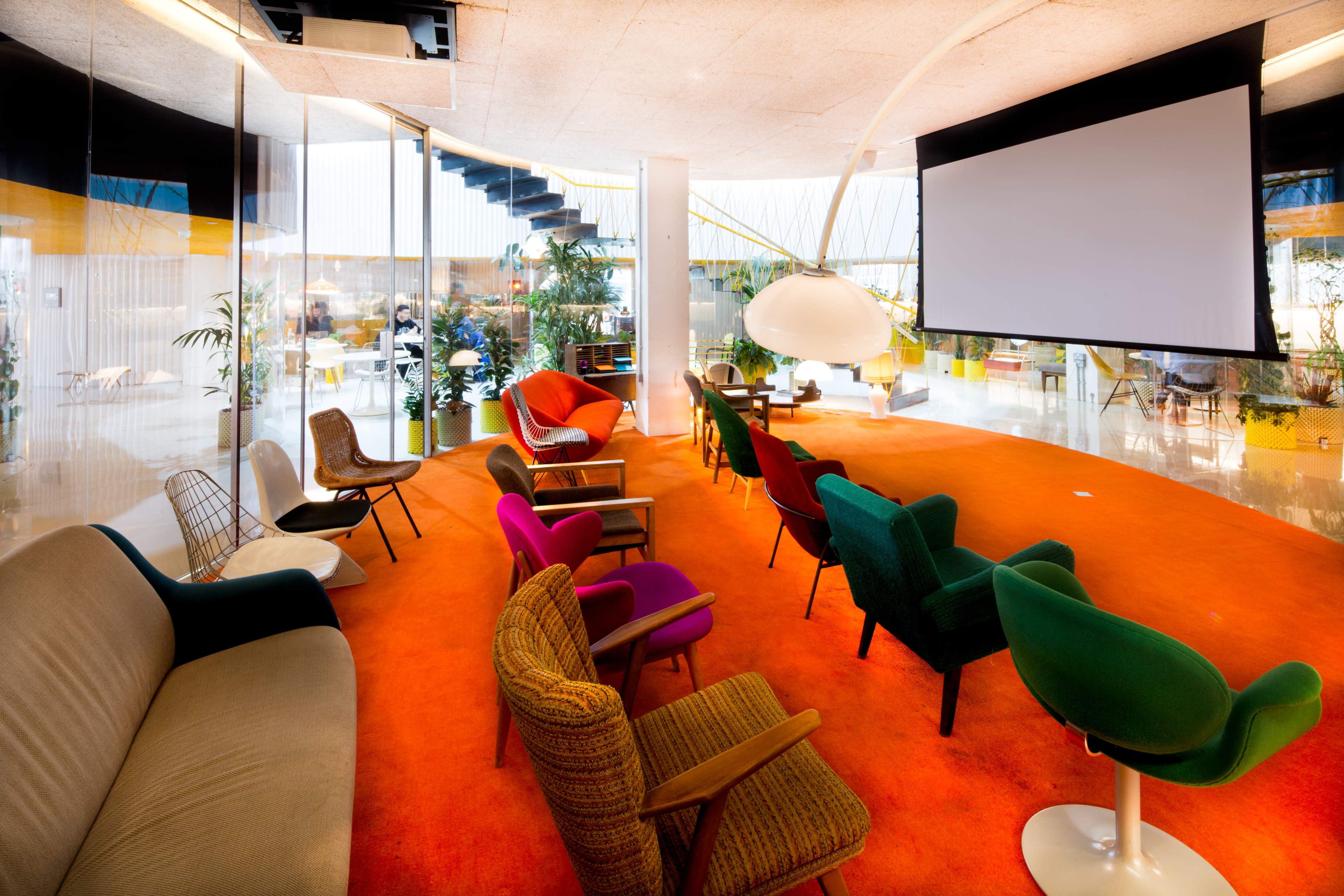 A lounge area features a variety of colorful chairs arranged around a large screen, with a vibrant orange carpet and plants in the background.