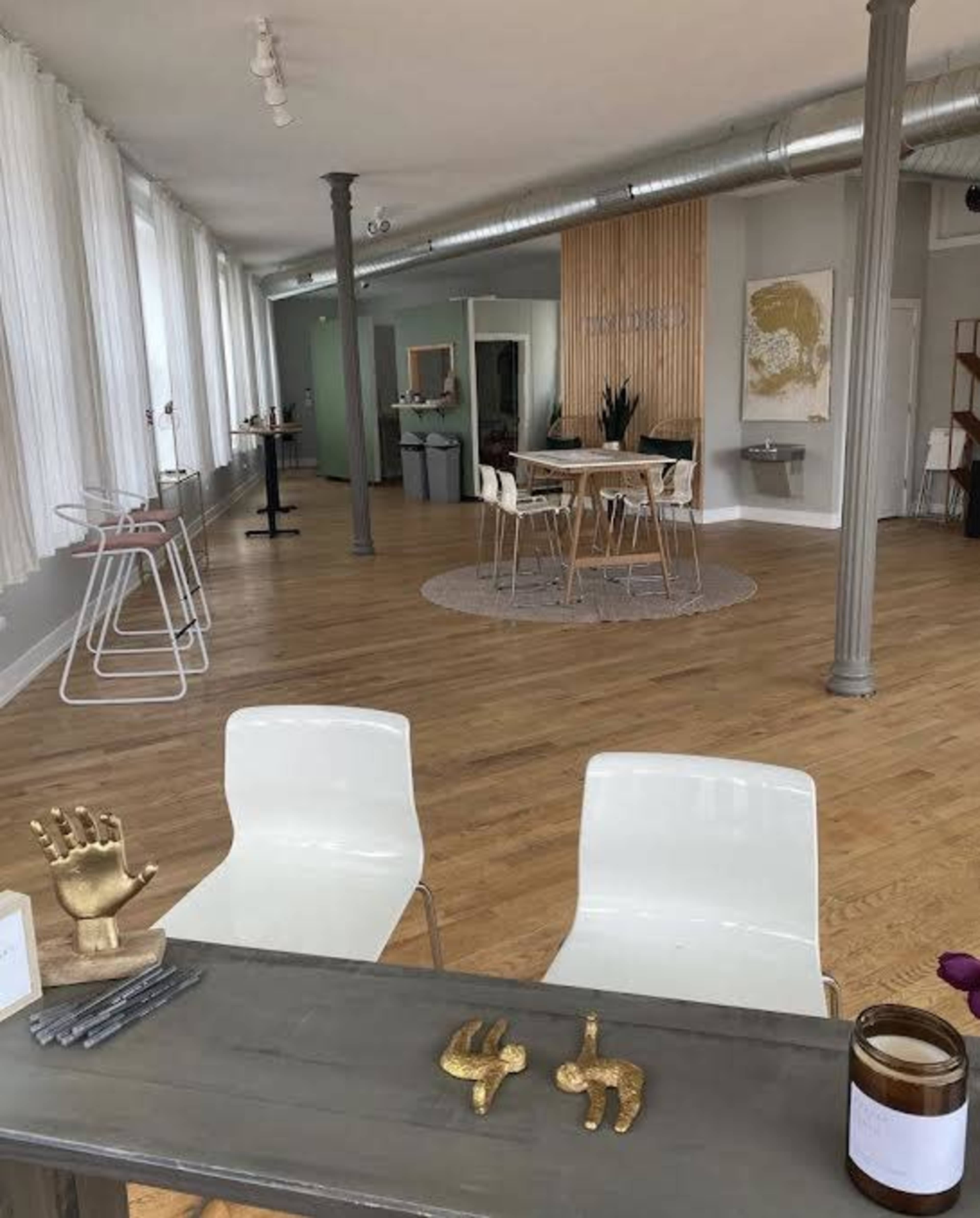 A spacious interior with wooden flooring, featuring a seating area with two white chairs, a table with decorative objects, and dining tables arranged in a well-lit space.