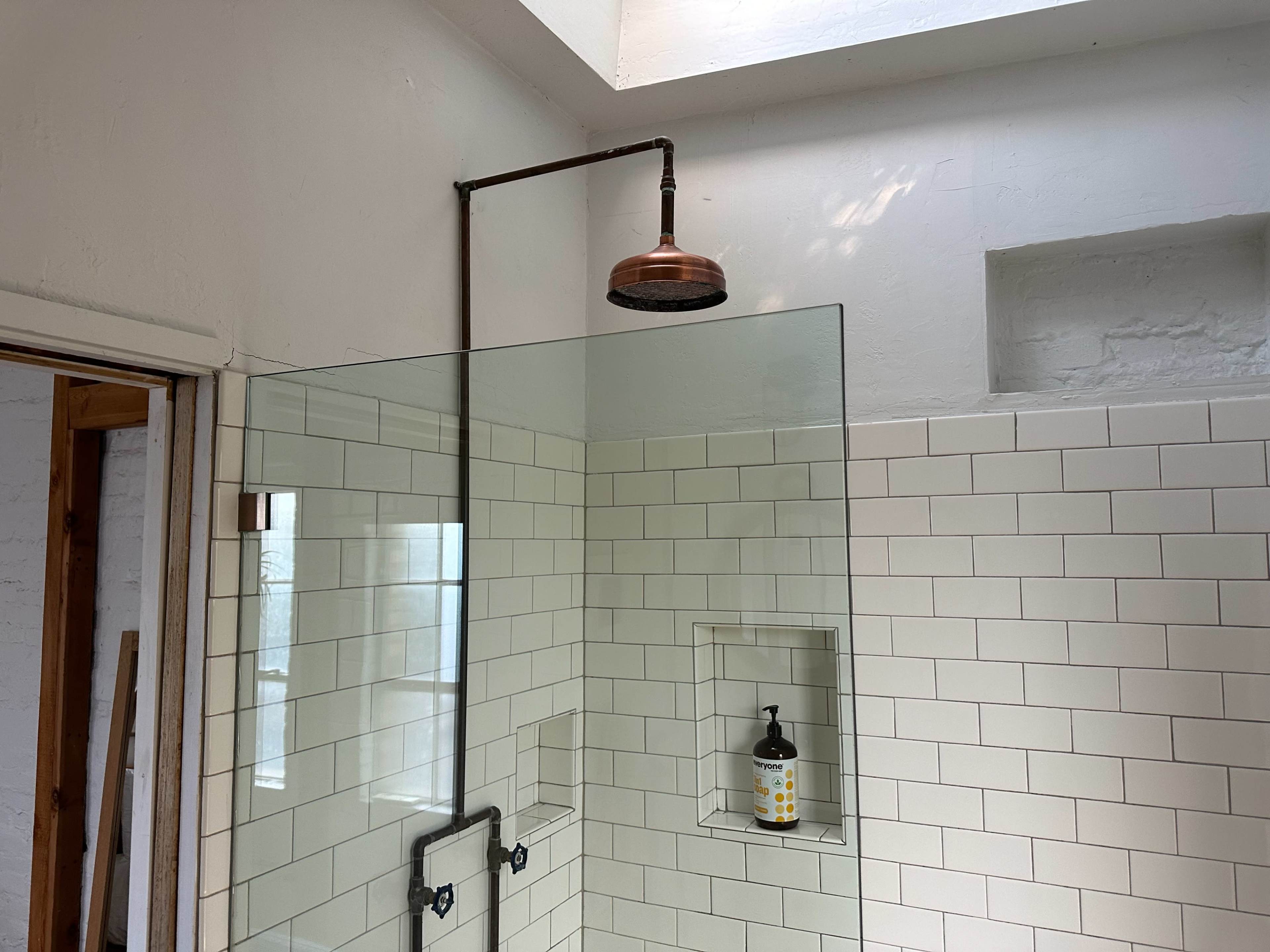 A shower area with a copper showerhead and a glass partition, featuring white tiled walls and a skylight above.
