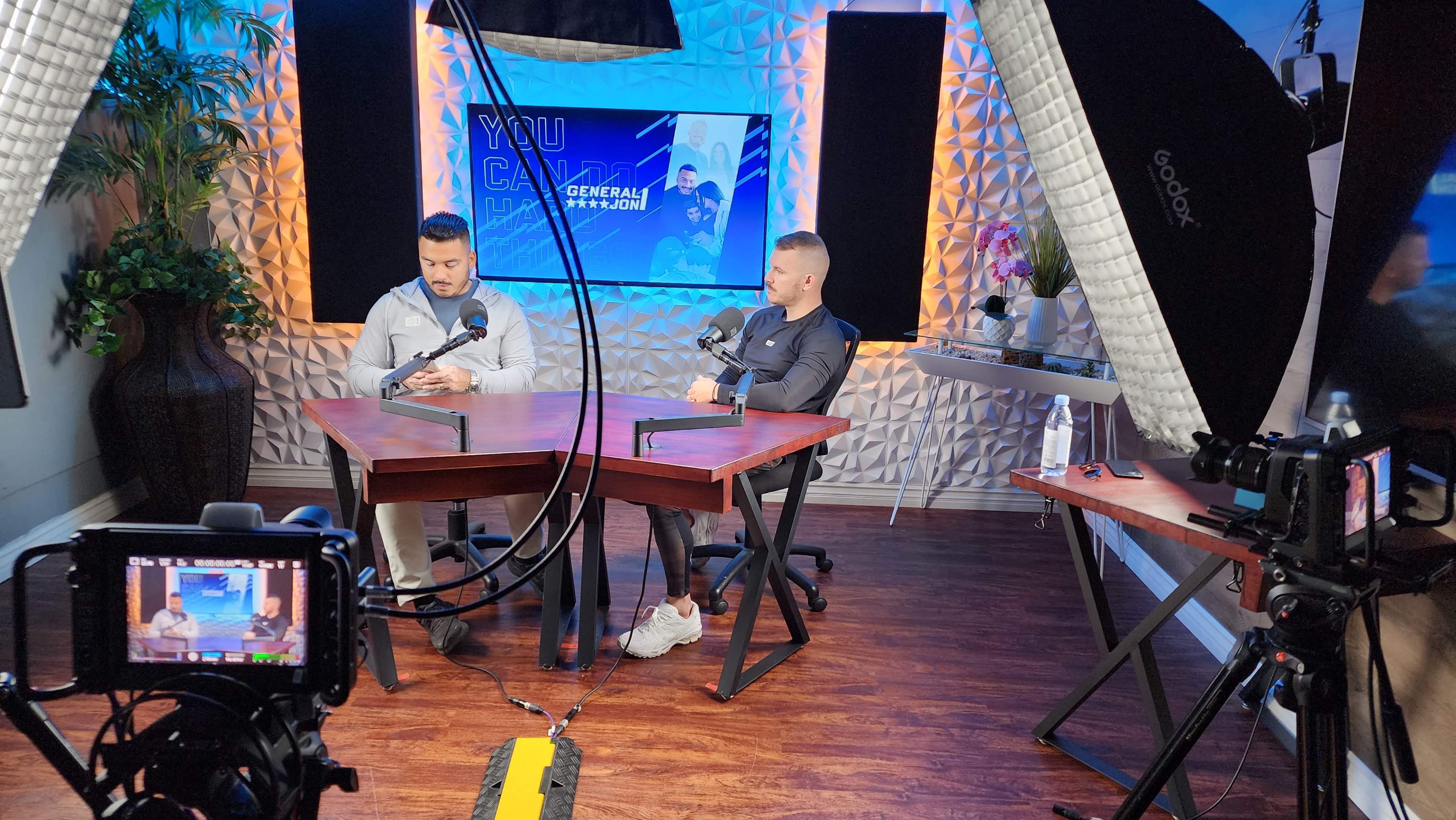 Two men are seated at a wooden table with microphones, surrounded by studio lights and a backdrop displaying the words "You Can" on a screen.