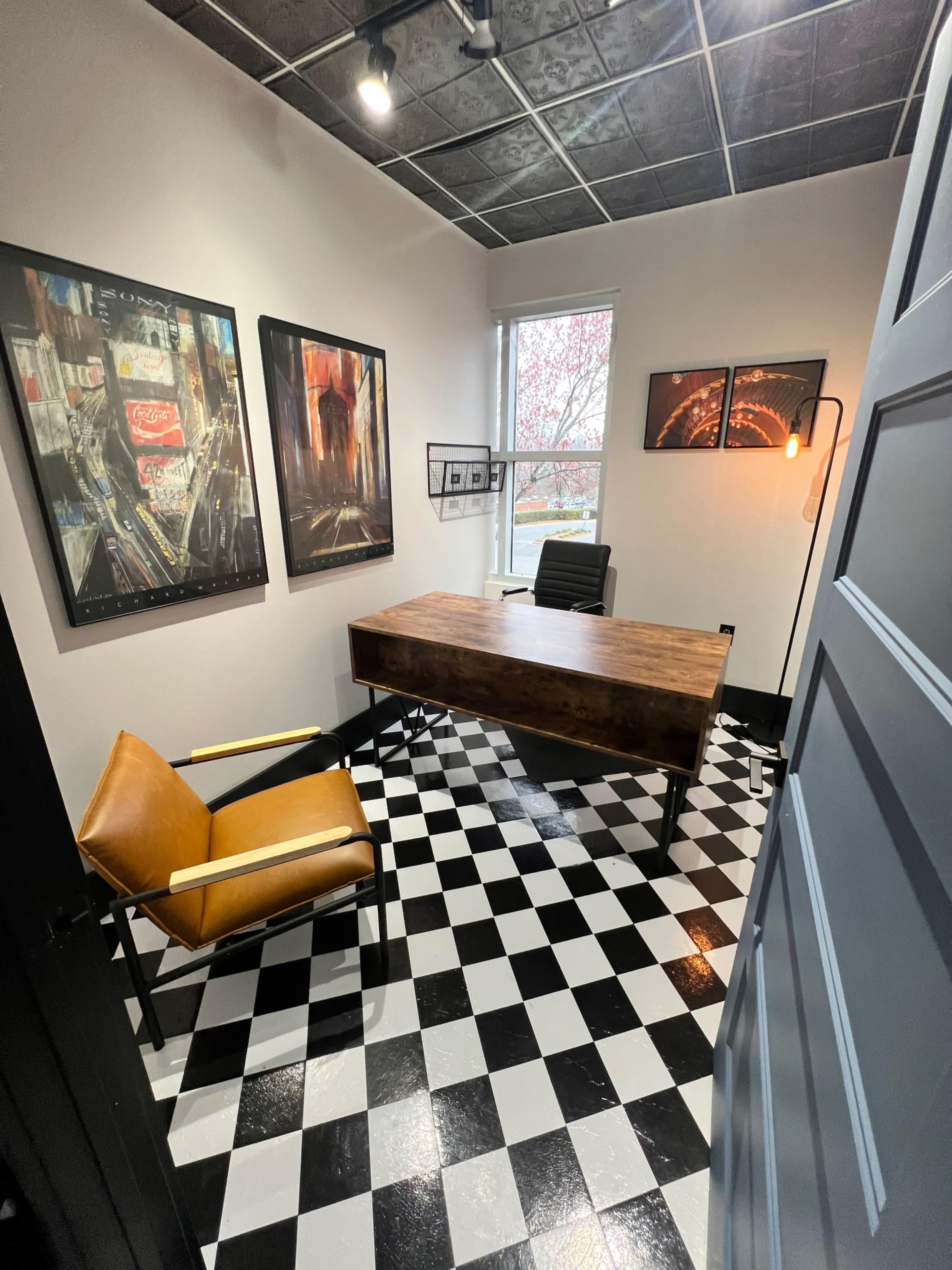 The image shows a modern office space featuring a wooden desk, a black chair, and a yellow armchair, with black and white checkered flooring and artwork on the walls.