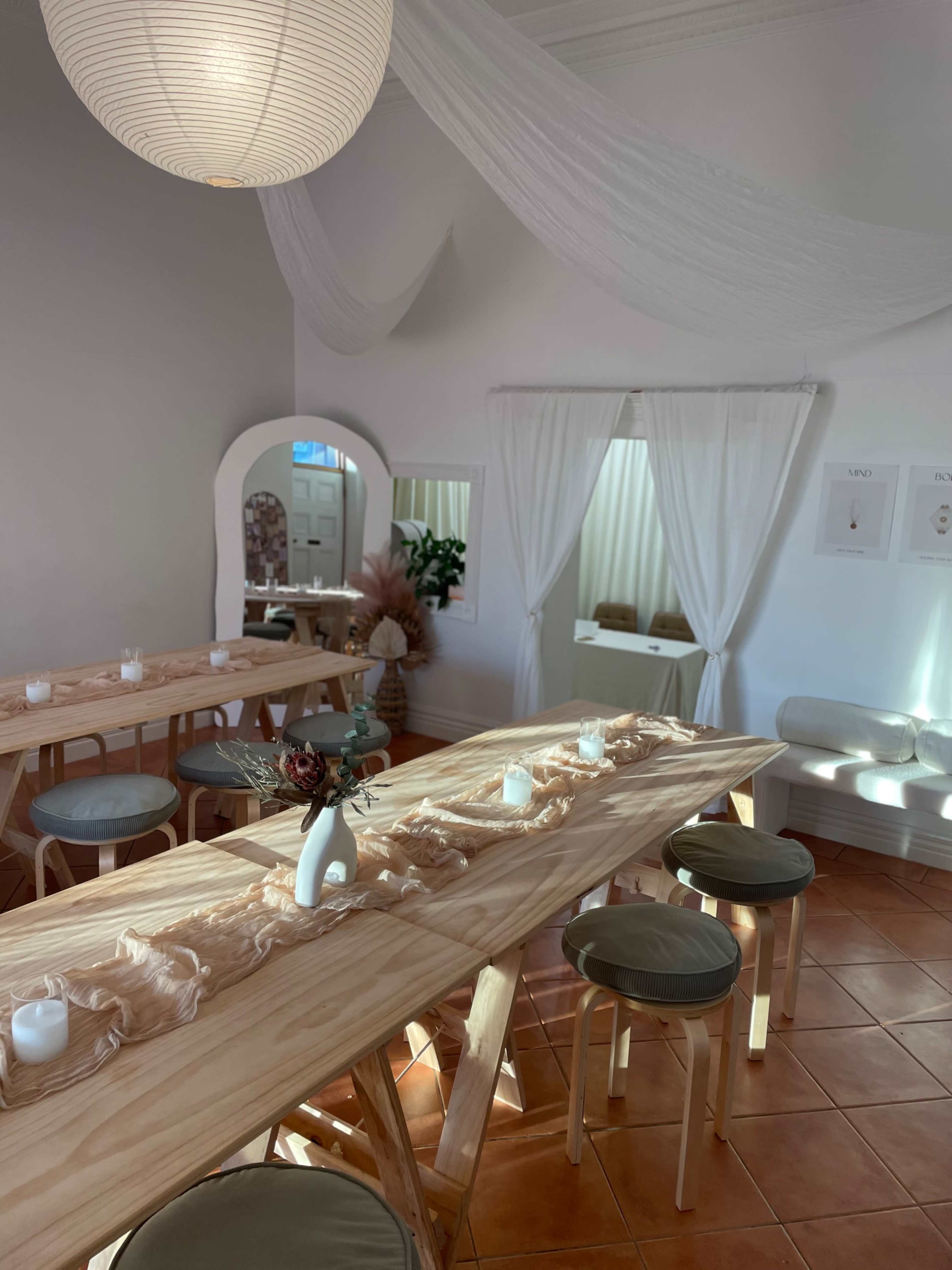 The image shows a softly lit dining area with wooden tables, minimalist decor, and a curtain that separates a small nook.