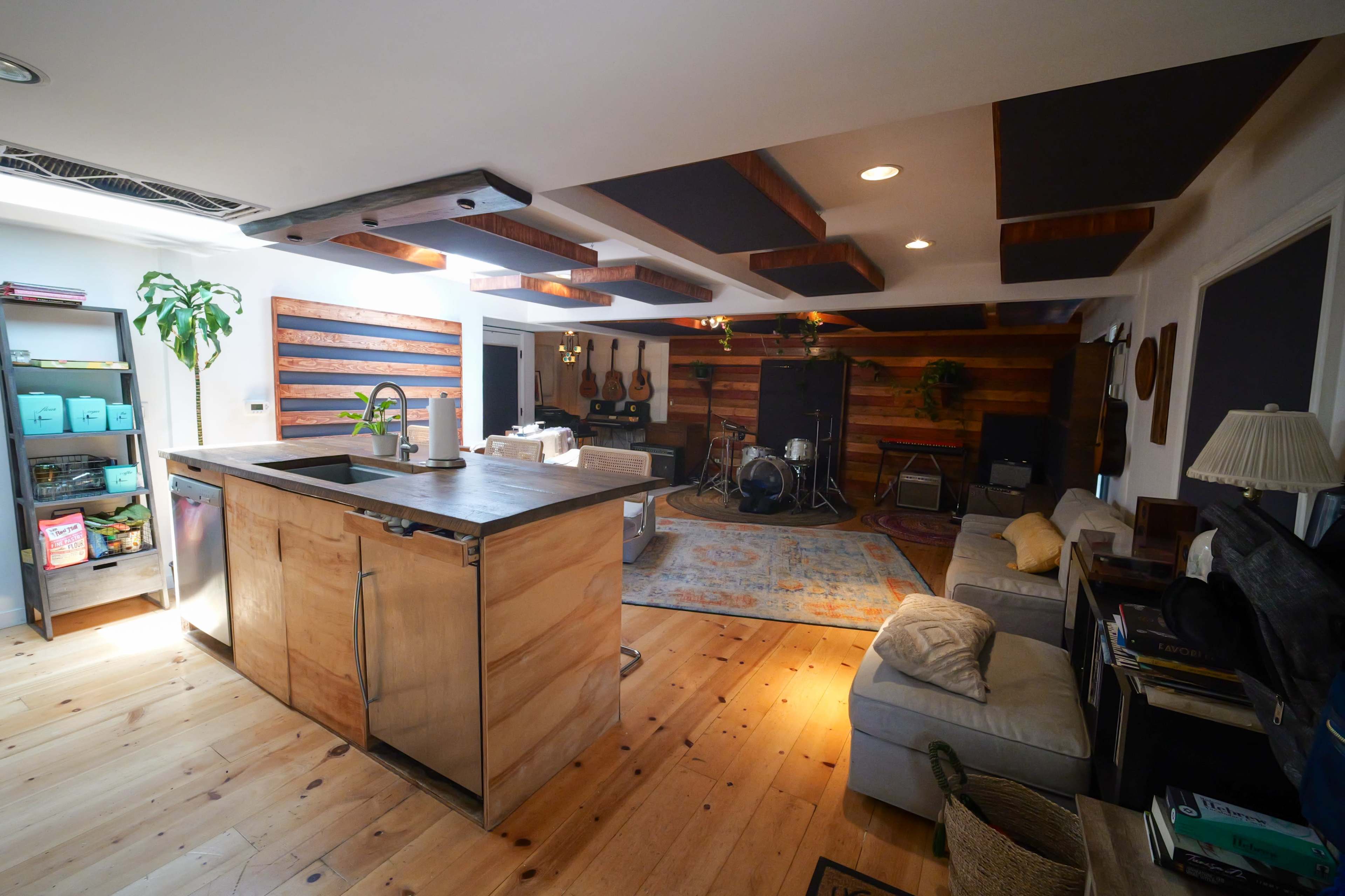 A spacious room featuring a kitchen area with a metal-topped island, wooden ceiling beams, a sitting area with a couch, and a corner set up for musical instruments.