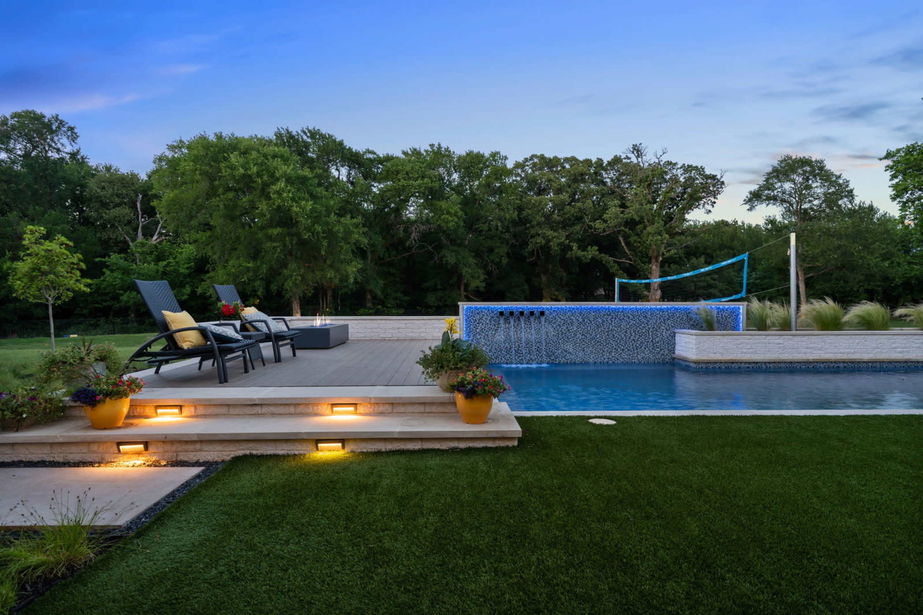 A backyard pool area with a modern infinity edge pool, outdoor seating, and illuminated stairs surrounded by greenery.