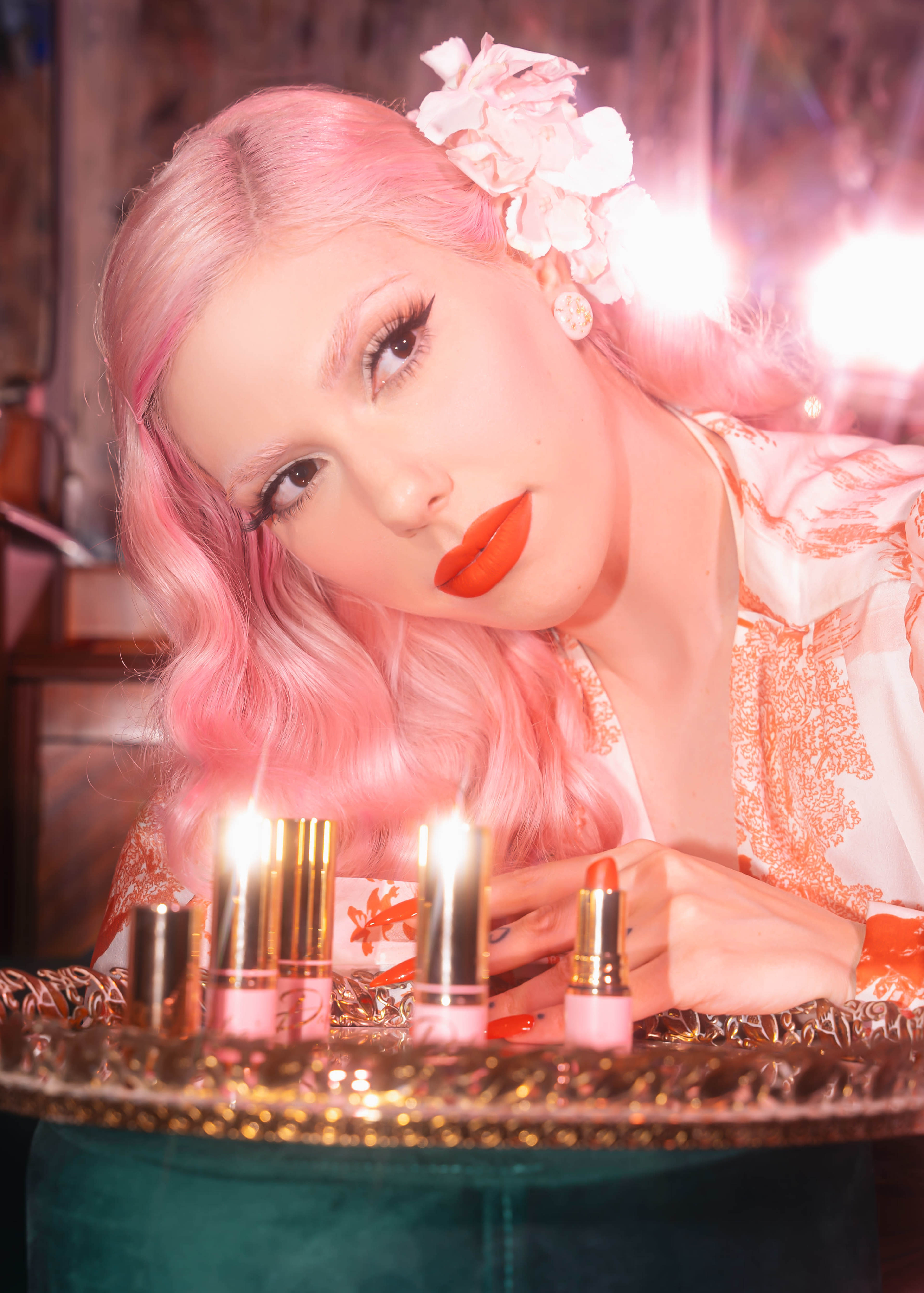 A woman with pink hair and bright red lipstick poses with several lipstick tubes arranged on a table in front of her, illuminated by soft lights behind her.