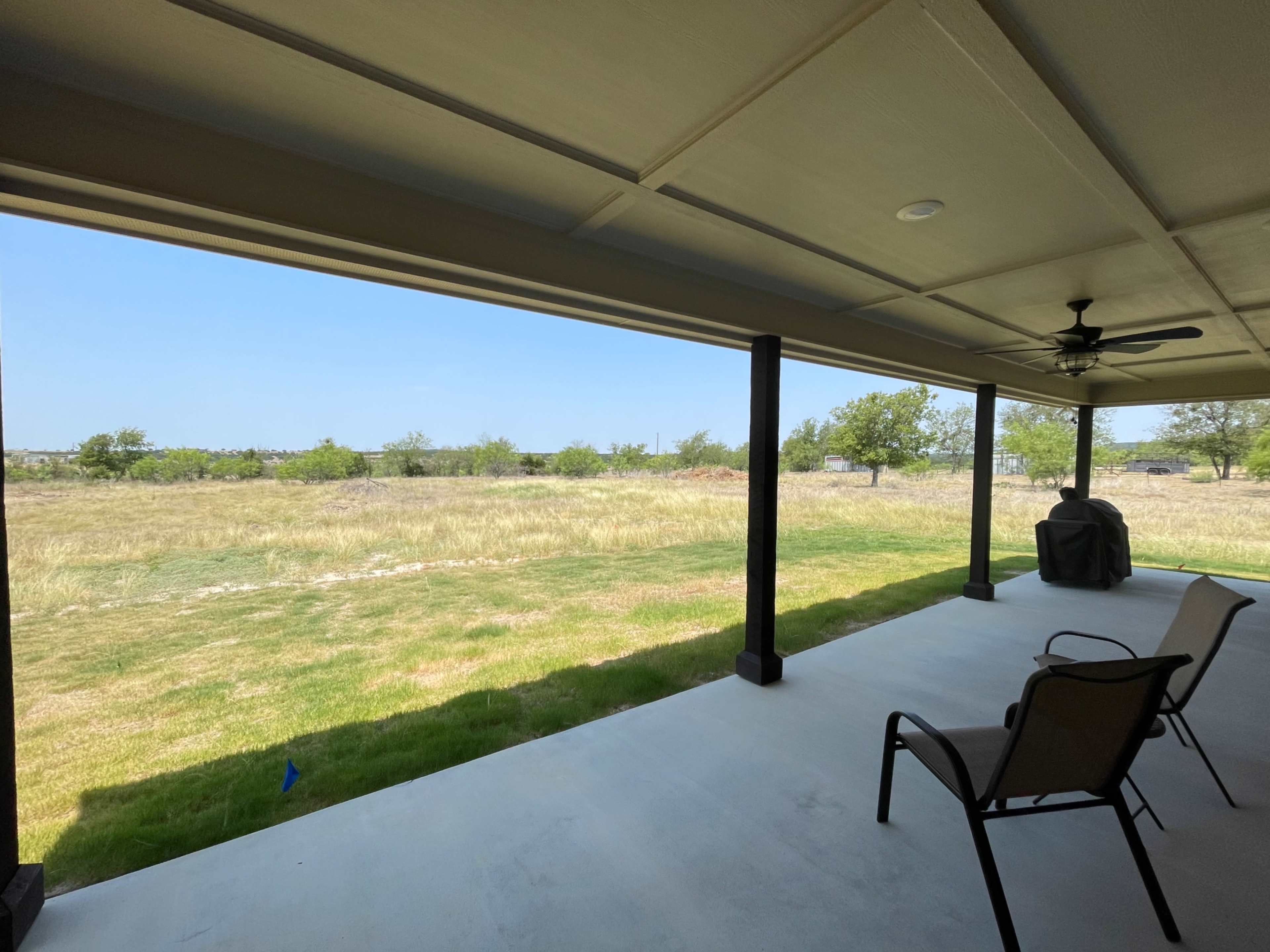 A covered patio with two chairs overlooking a grassy field and distant trees under a clear blue sky.