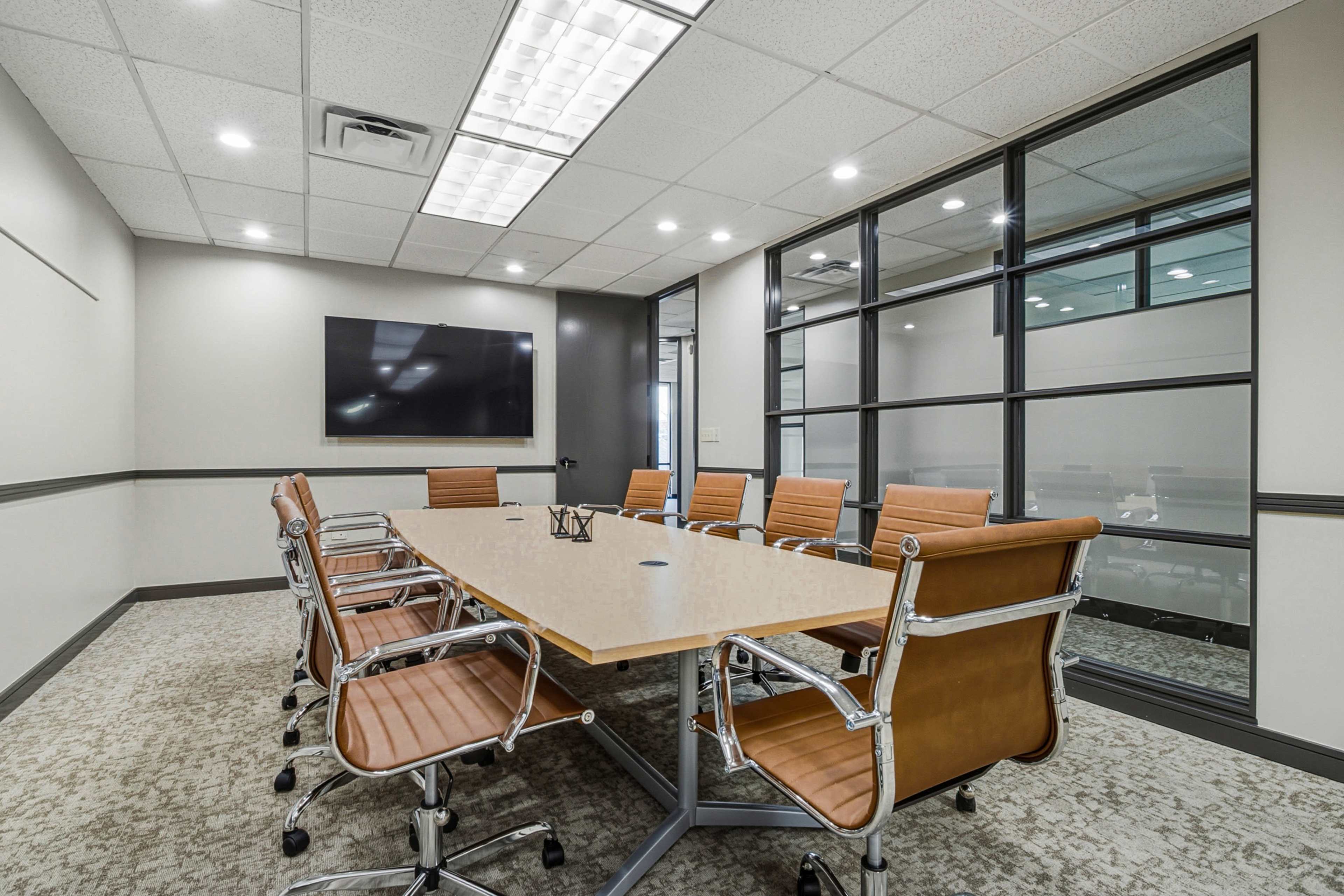 A modern conference room features a long wooden table surrounded by several tan leather chairs, with a large screen mounted on the wall and glass partitions.